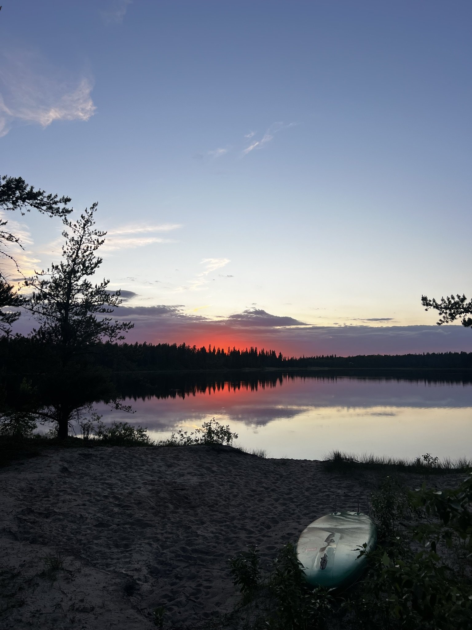 A tranquil lakeside scene at sunset with a calm water reflection, dark trees along the horizon, and a boat resting on the sandy shore.