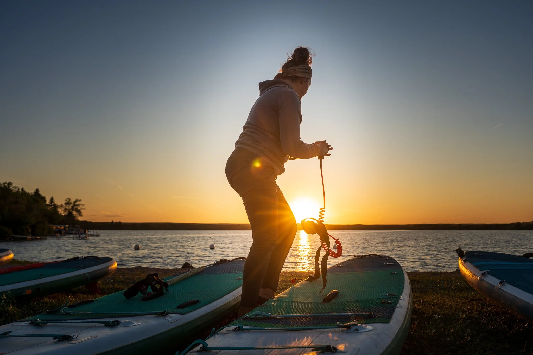 A person standing on a paddleboard at sunset, lead by Back2Nature Wellness, with paddleboards on the shore and a body of water in the background.
