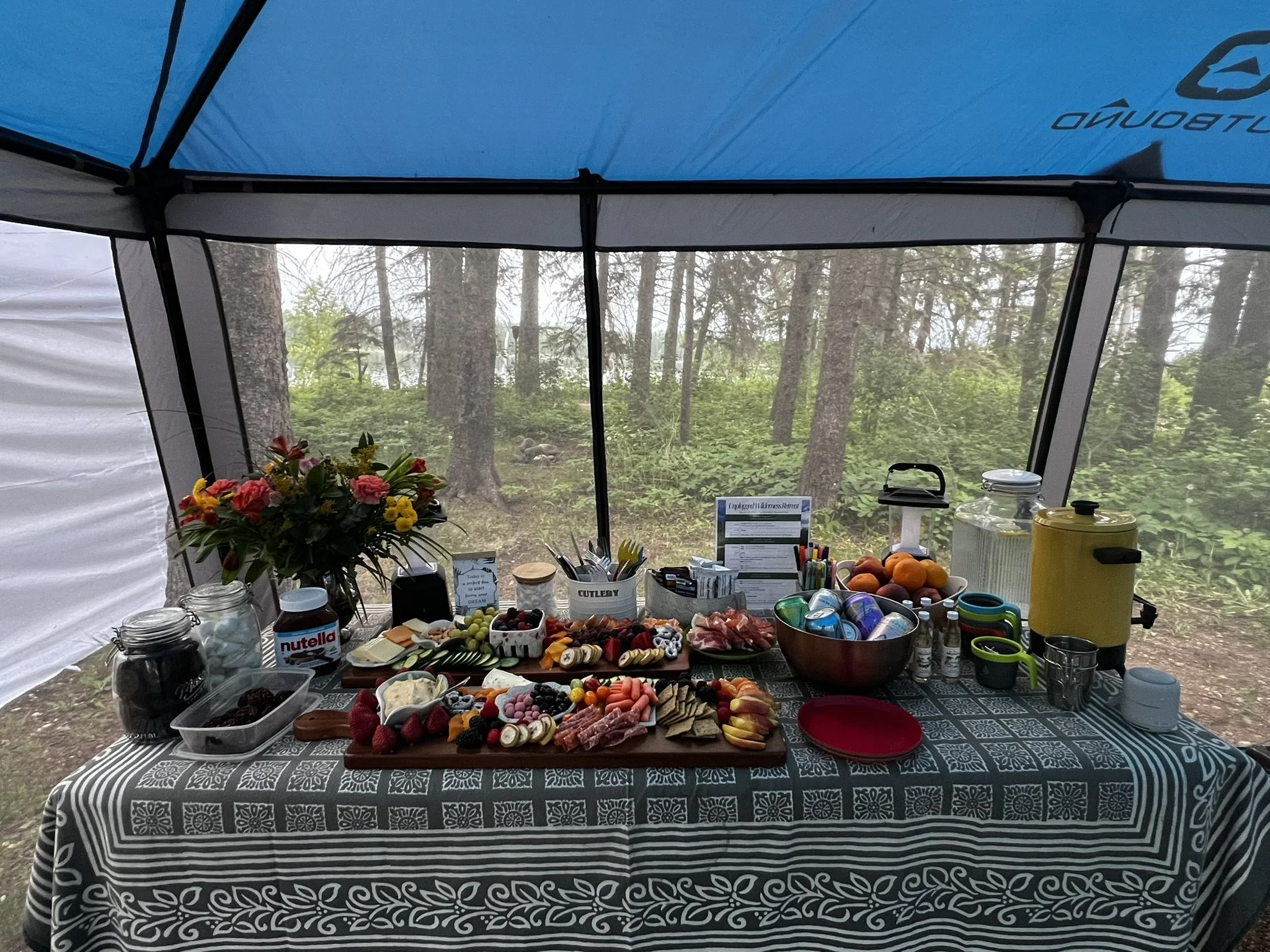 A picnic table set up outdoors in a forest with a variety of food items including fruits, cheeses, meats, and snacks, decorated with a bouquet of flowers, under a canopy tent.