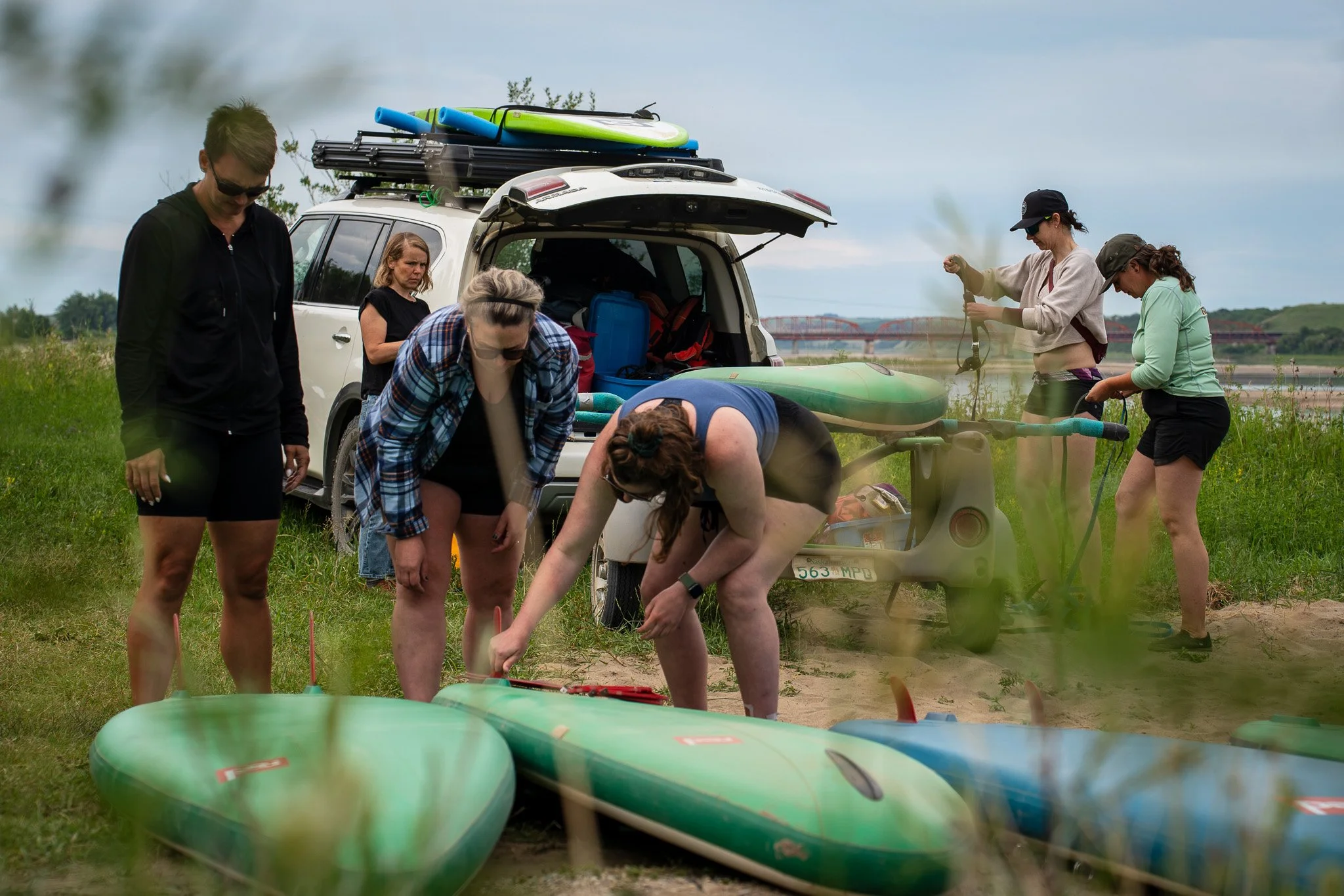 Group of people preparing paddleboards and gear at a grassy riverside, with a white vehicle in the background loaded with paddleboards.