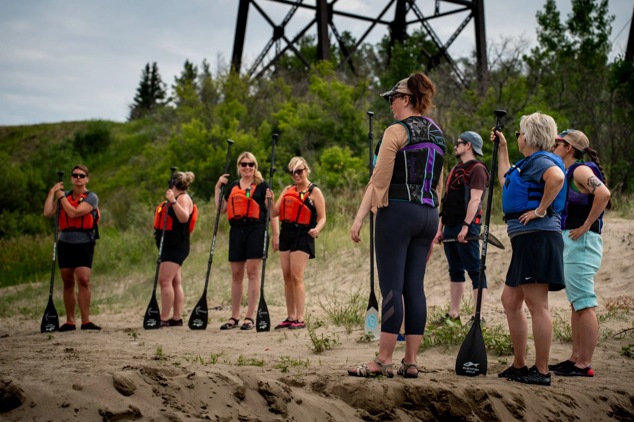Group of people preparing for a stand-up paddleboarding adventure on a sandy beach, holding paddles, with green trees and a power line tower in the background. Overnight Stand-up Paddleboard Camping Adventure