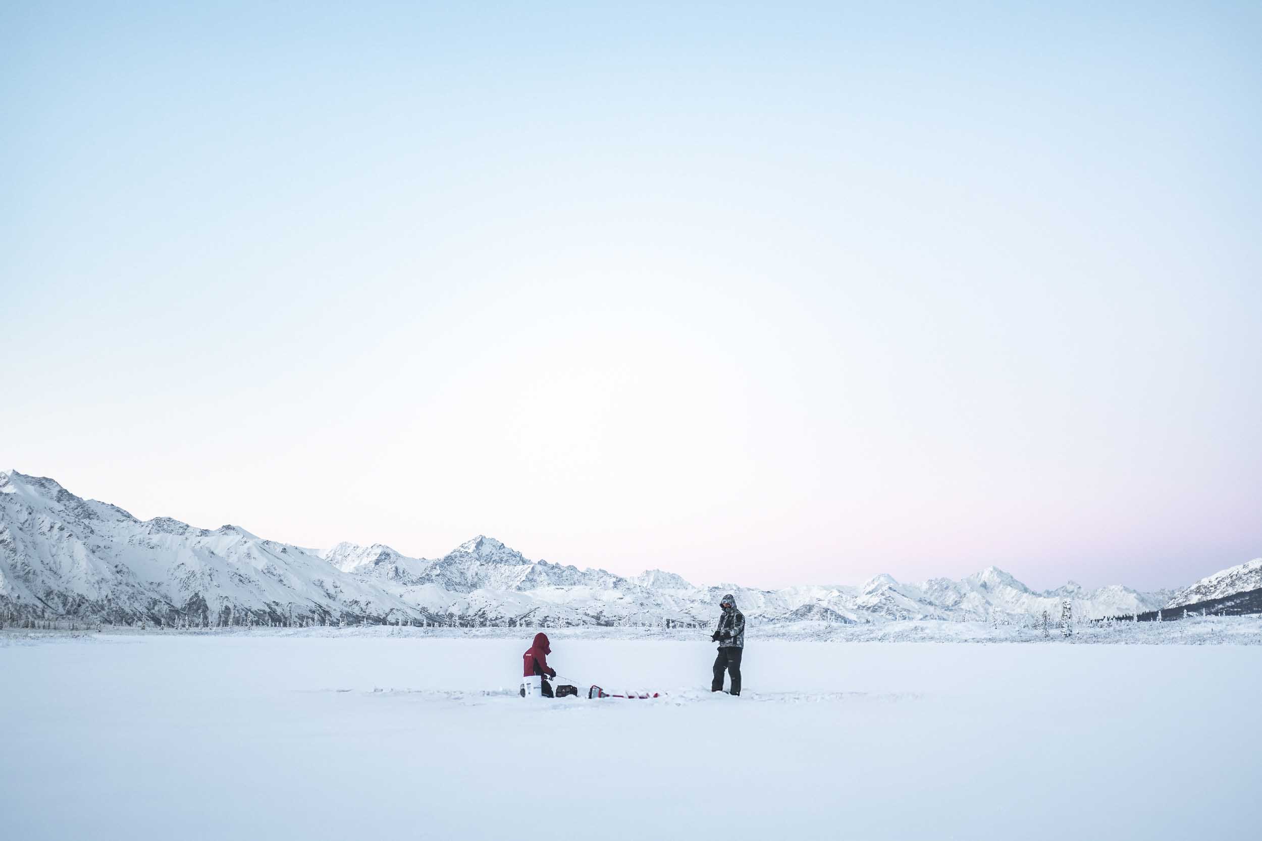 Wide shot of two people ice fishing on a frozen lake during a photo and video shoot in Alaska, showcasing adventure photography and commercial video production.