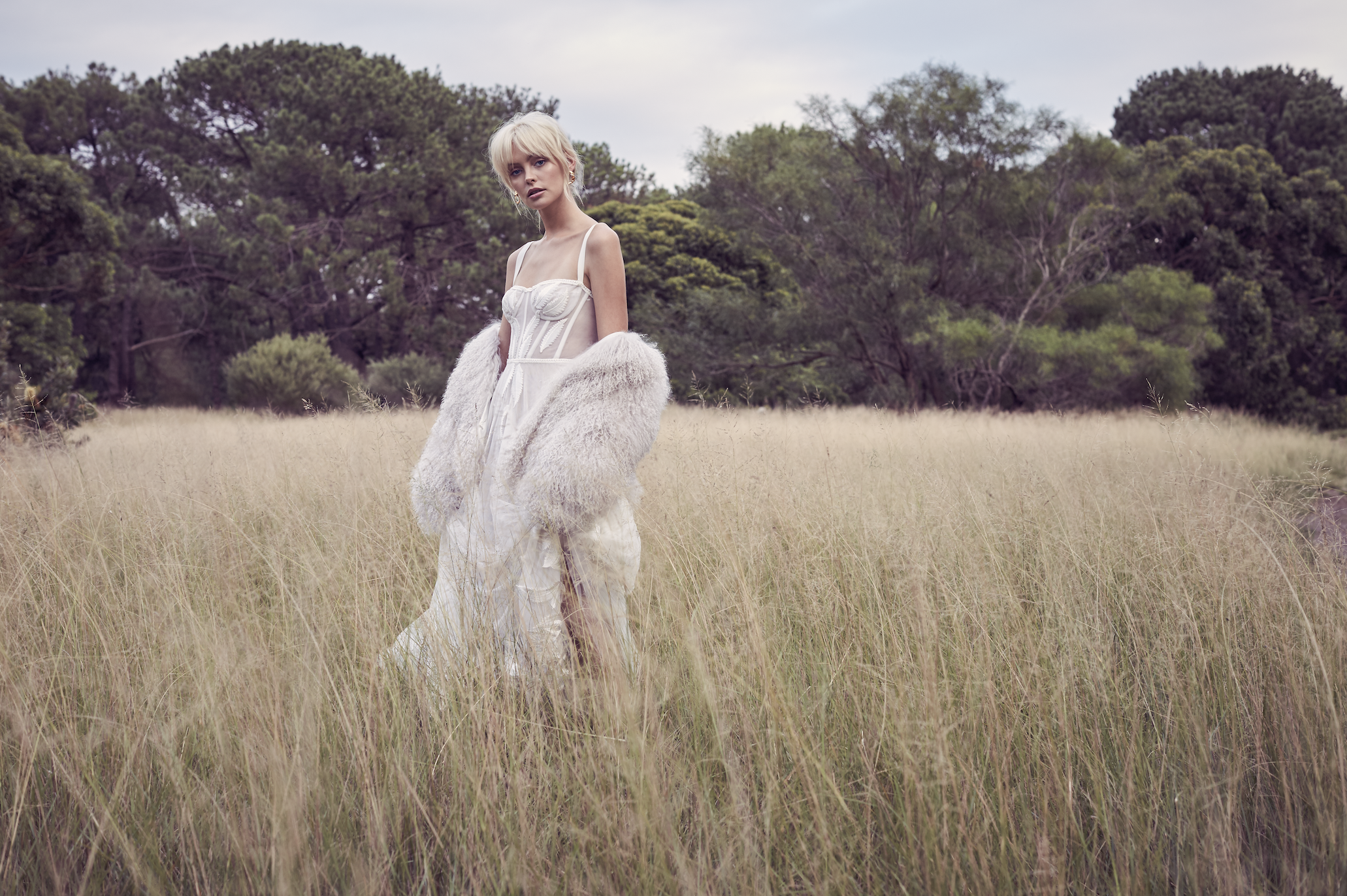 A woman in a white dress and fuzzy coat standing in a grassy field with trees in the background.