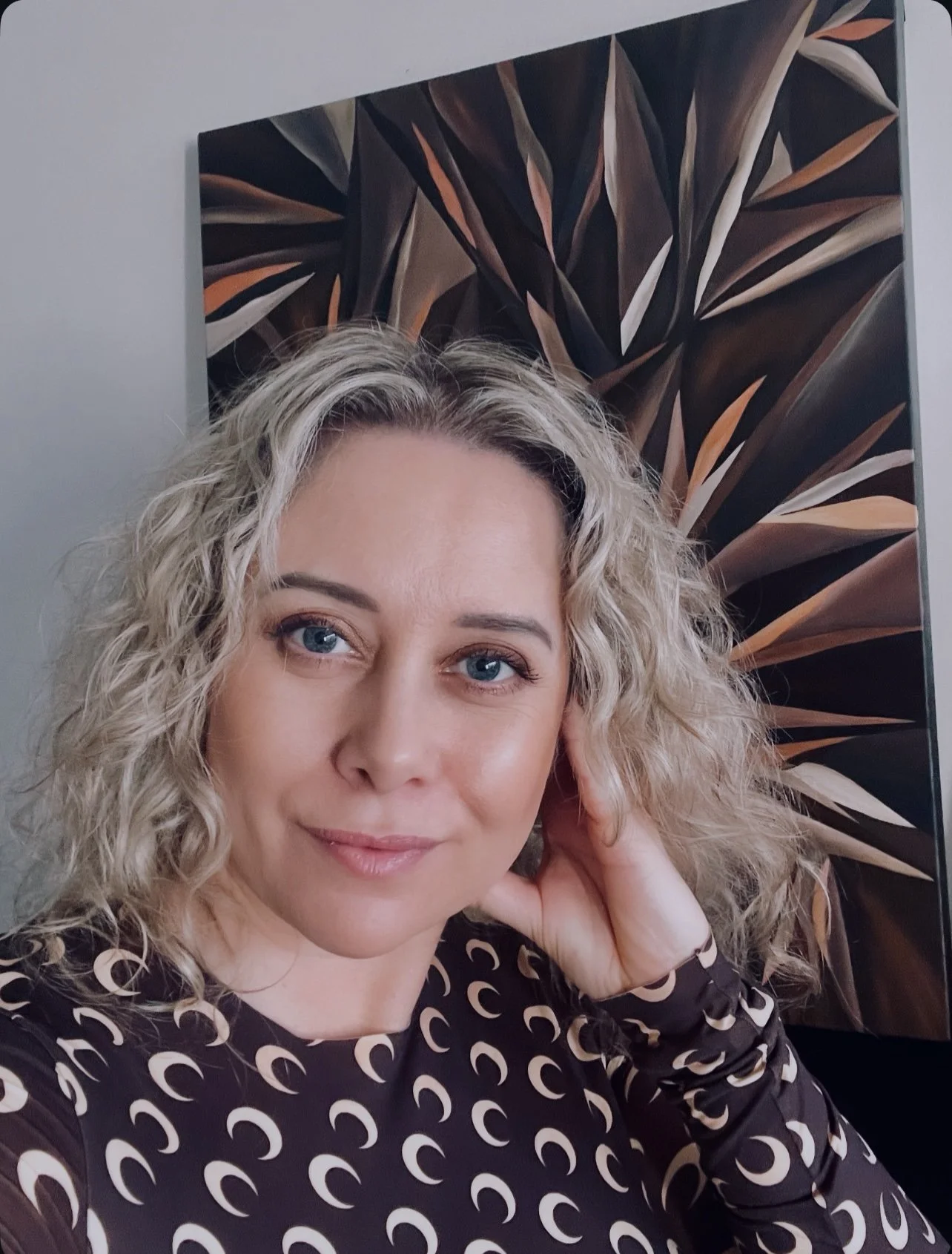 A woman with curly blonde hair, blue eyes, and light makeup smiling softly and resting her hand near her face, wearing a dark patterned top. Behind her is a large abstract artwork featuring dark and brown tones with leaf-like shapes.