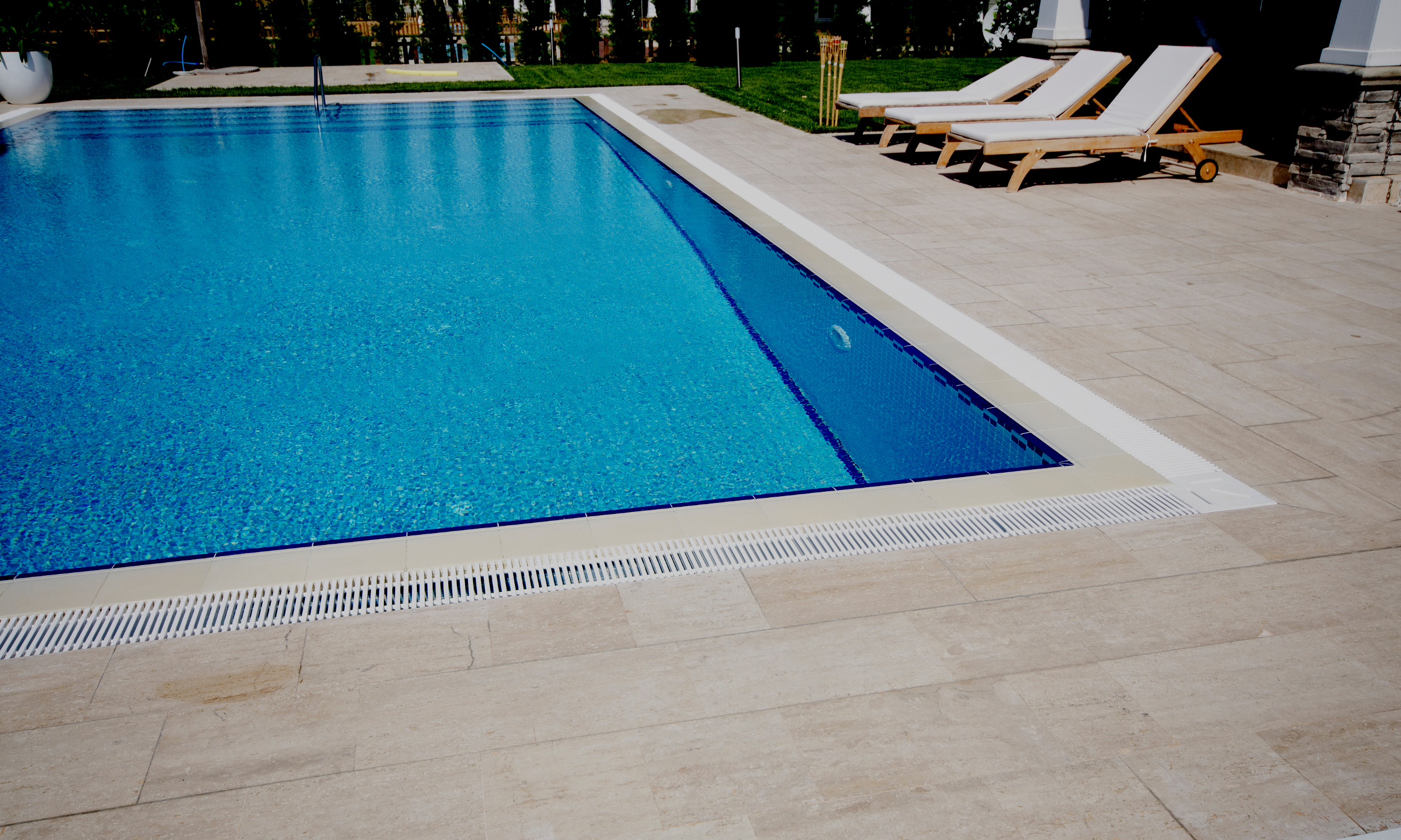 A backyard swimming pool with blue water, surrounded by beige stone tiles, four lounge chairs with white cushions, and a landscaped grassy area with trees and bushes.
