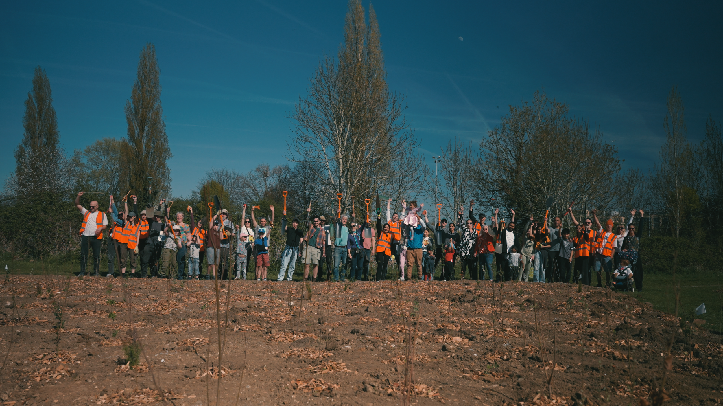 A large group of people wearing orange safety vests standing on dirt ground outdoors, holding shovels upright, with trees and a clear blue sky in the background.