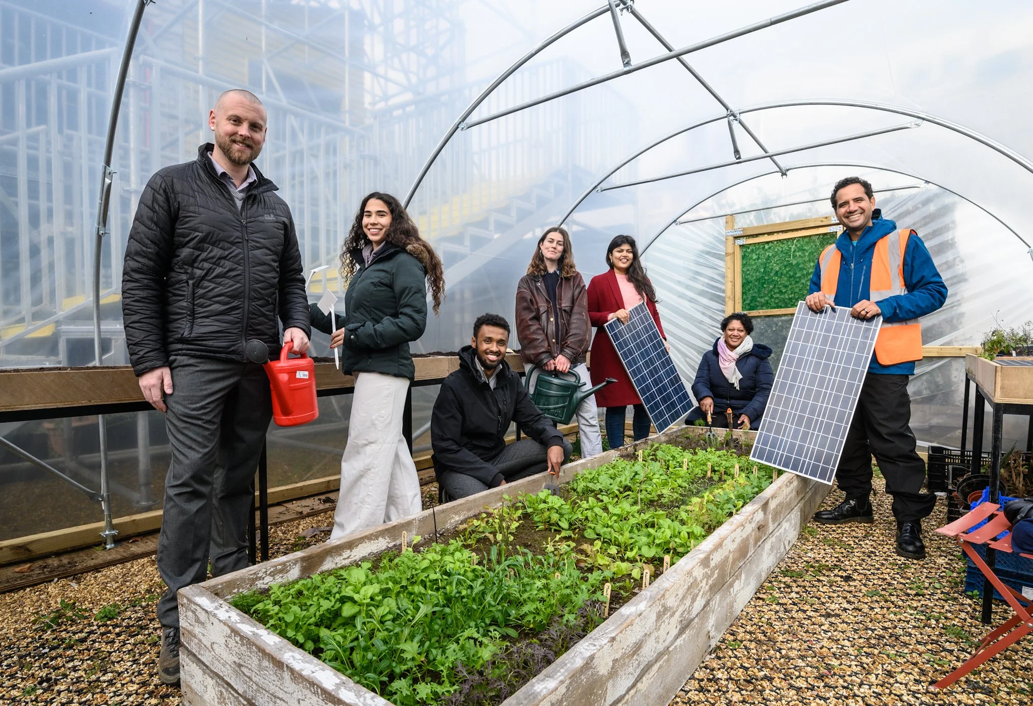 Group of people in a greenhouse with solar panels, gardening tools, and plants, smiling at the camera.