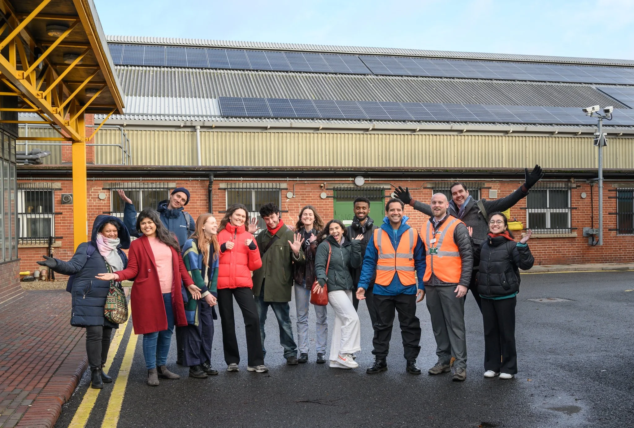 Group of diverse people smiling and posing outdoors in front of a building with solar panels, some wearing safety vests.