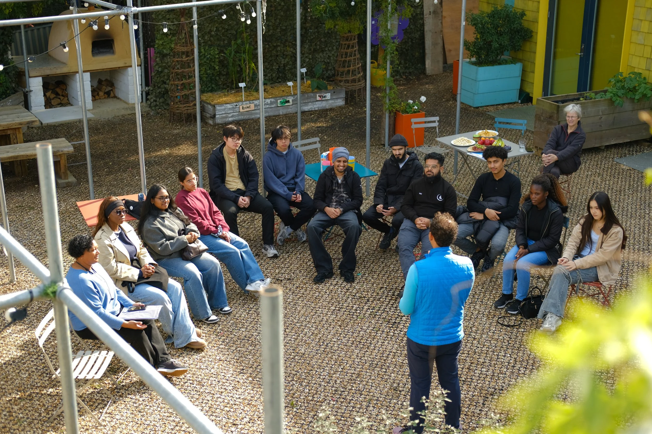 A group of people seated in a semi-circle outdoors, listening to a person standing and speaking. There are tables with food and drinks behind them, and a woman observing from a distance.