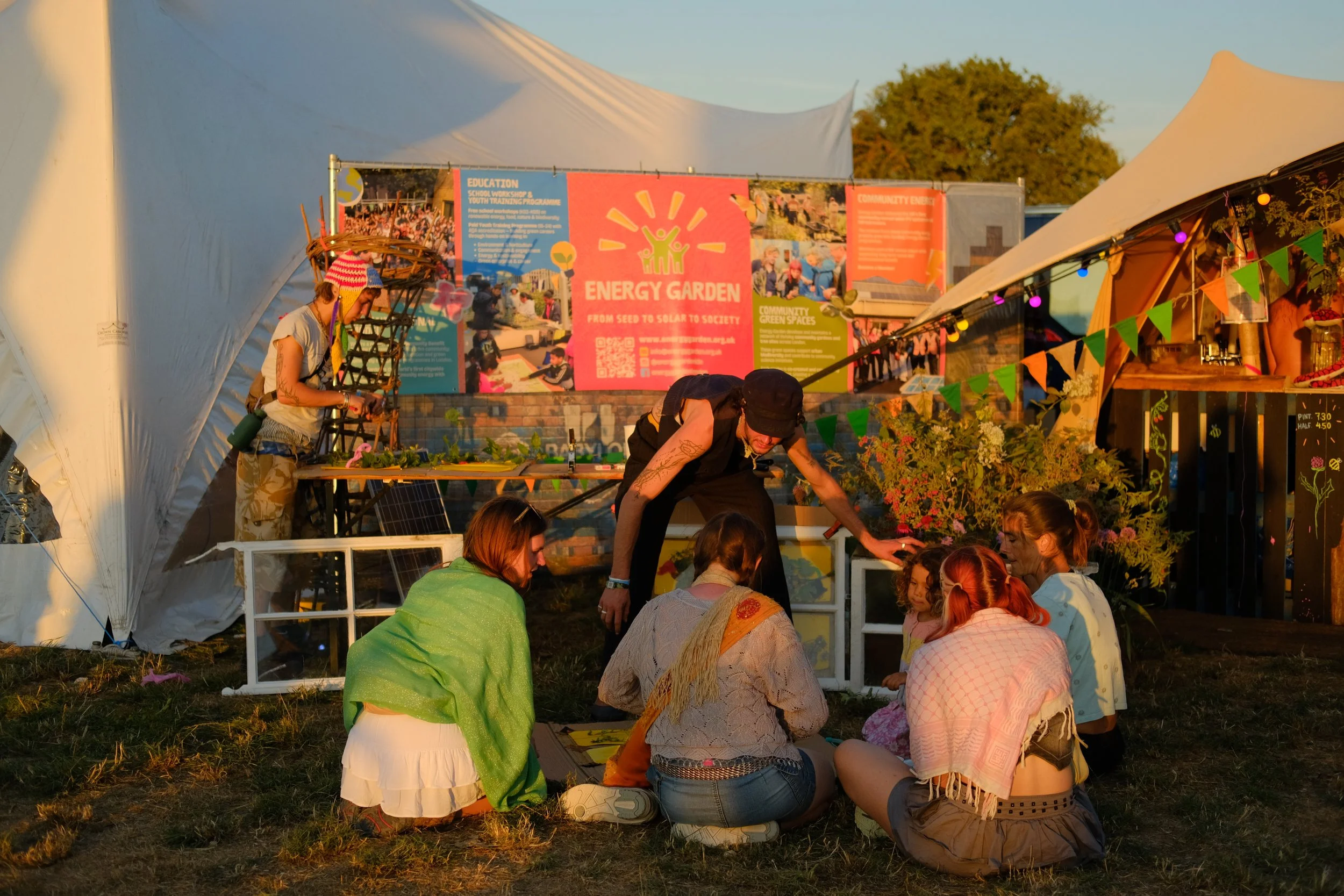 People gathered at an outdoor event under tents, with a large pink sign that readsEnergy Garden. Notes and various plants are displayed nearby, and the group appears to be engaged in a discussion or workshop during sunset.