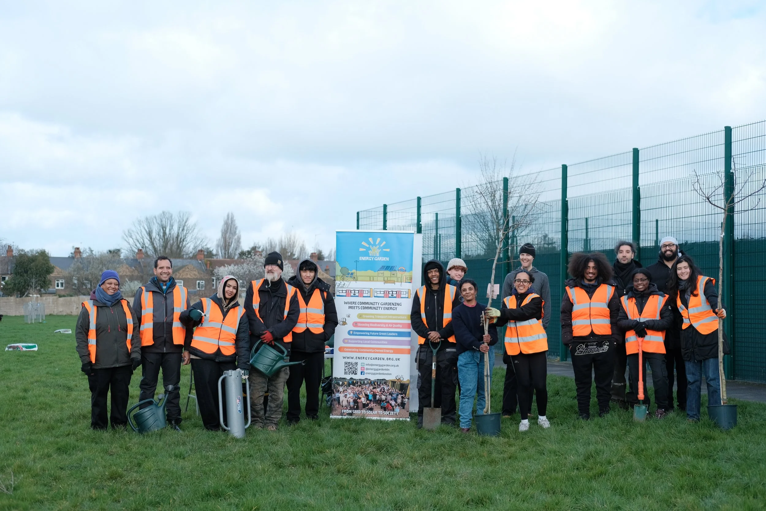 Group of people wearing orange safety vests participating in a community gardening event in an open grassy area, holding plant saplings and gardening tools, with a banner promoting energy and community garden initiatives.