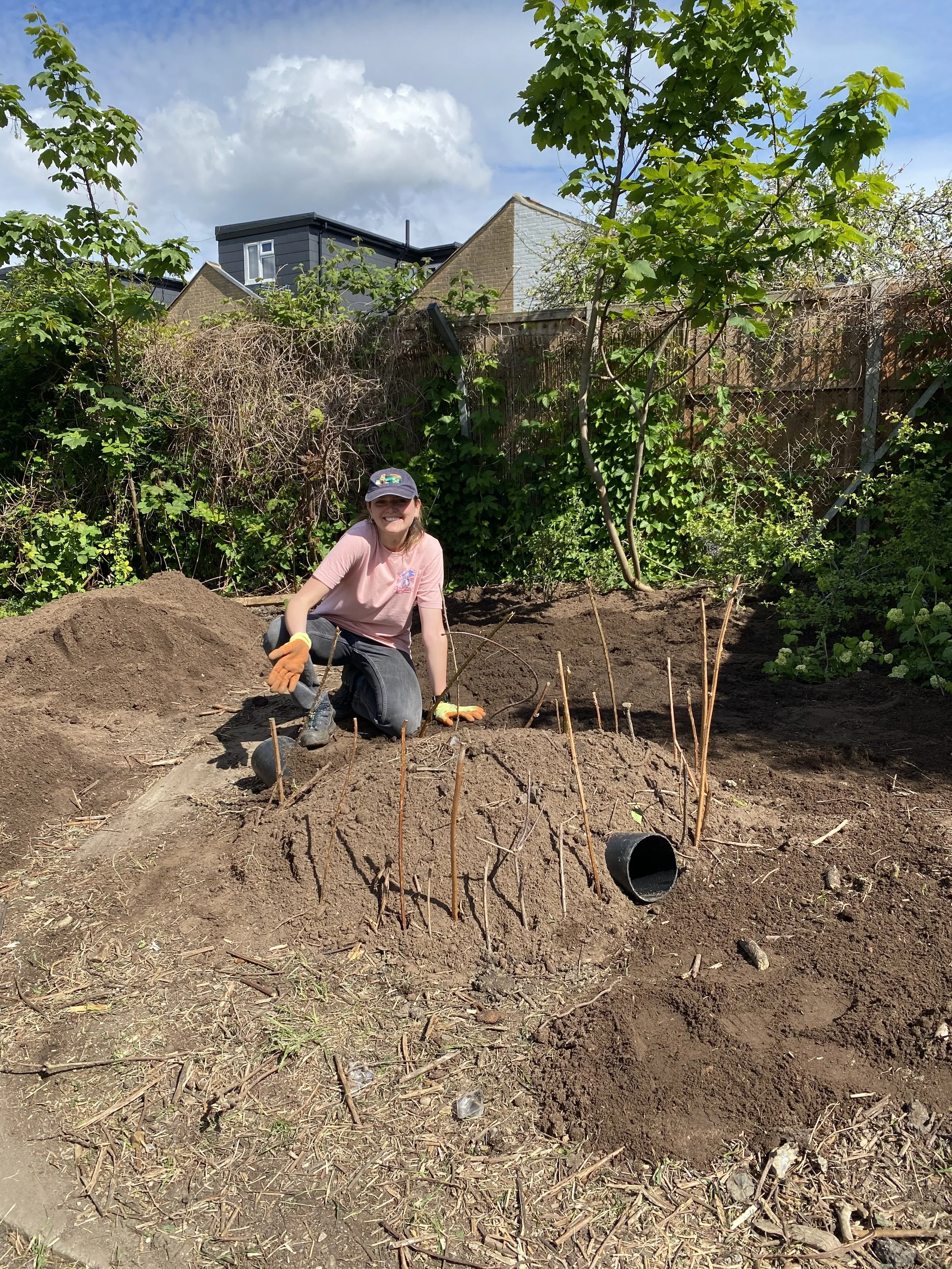 A young girl with a cap and gloves kneeling on the ground, smiling, in a garden with dirt mound, small sticks, and a black pipe, with trees and houses in the background.