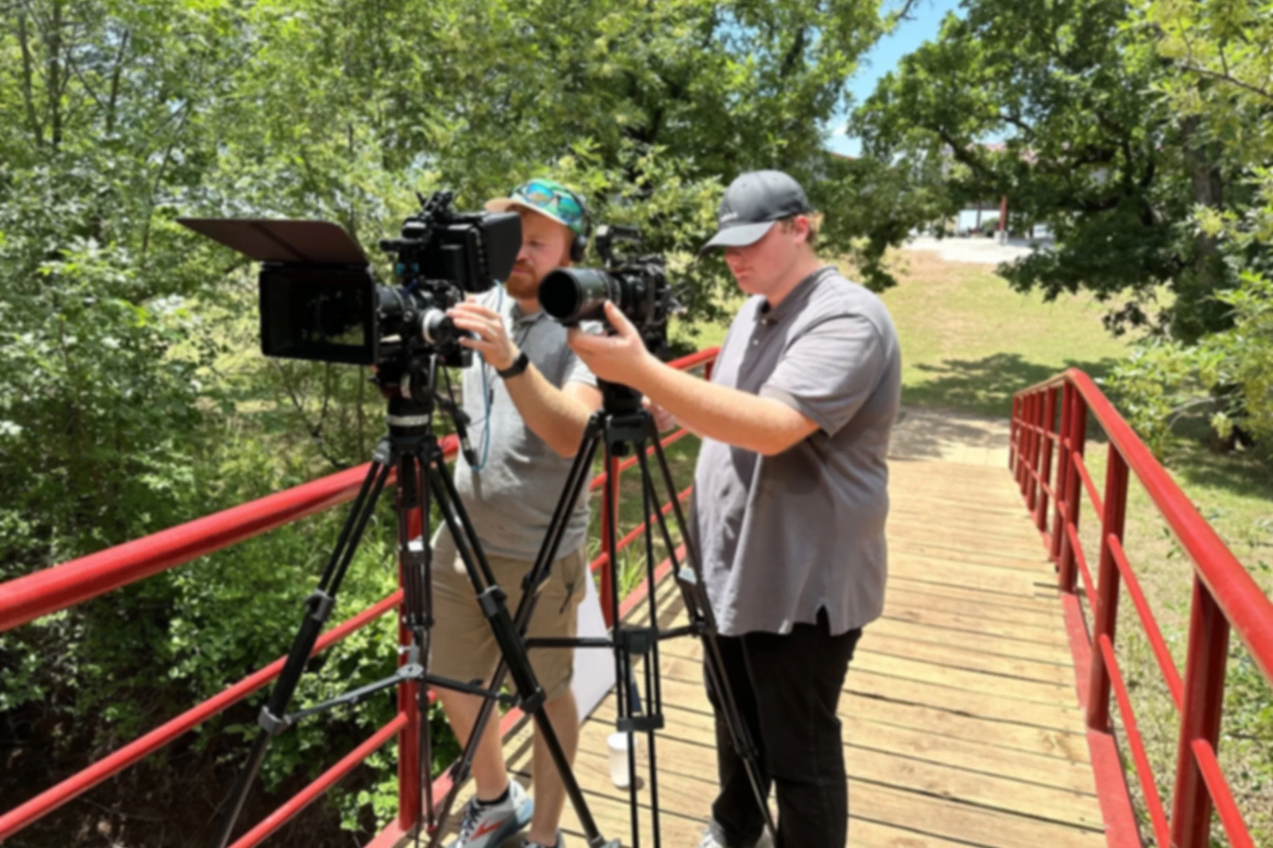 Two men filming with professional cameras on a wooden bridge surrounded by trees on a sunny day.