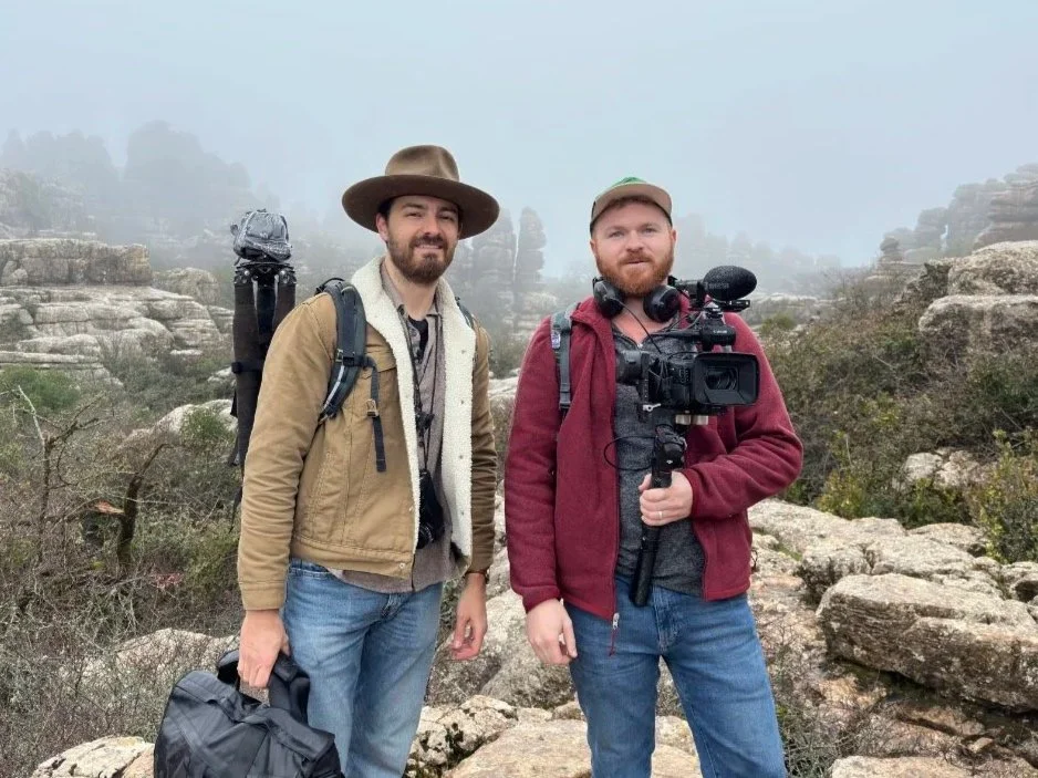 Two men standing outdoors on rocky terrain with a foggy background. One is holding a camera, and the other has a backpack and a bag. They are dressed casually for outdoor activities.