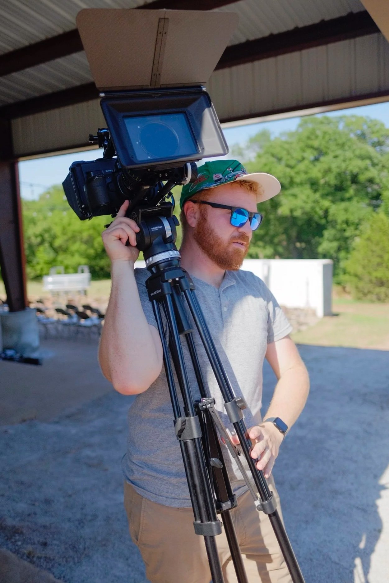 A man with red hair and a beard wearing sunglasses, a green cap, a light gray shirt, and khaki shorts, walking outdoors under a covered seating area moving a professional video camera and tripod on his shoulder.