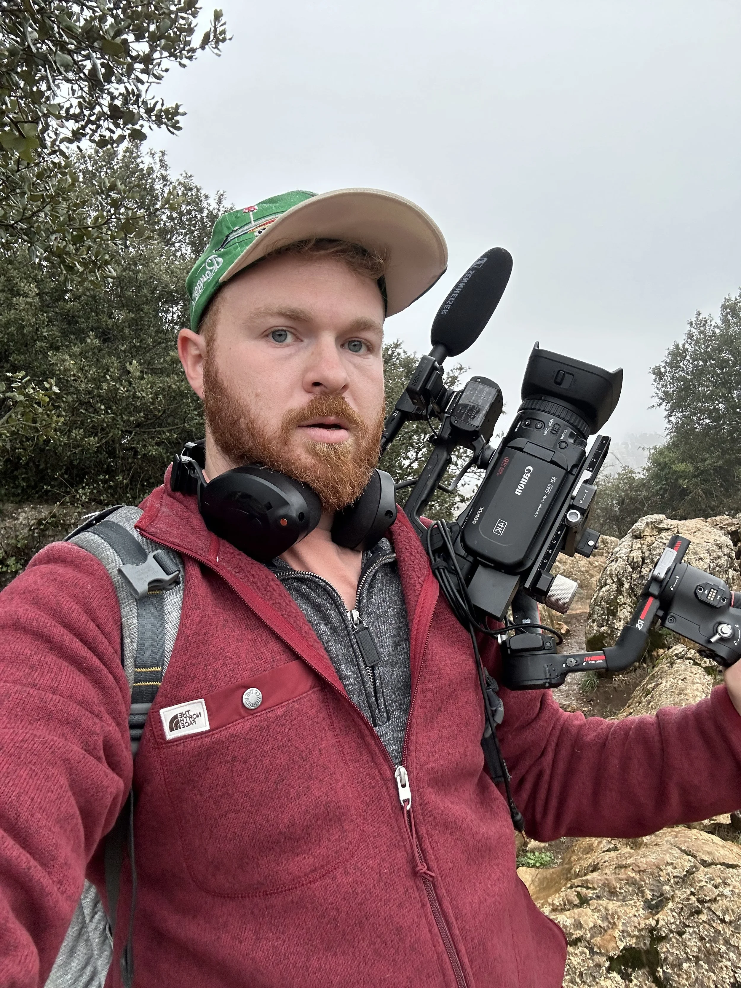 A man with a red beard and blue eyes wearing a beige cap, red jacket, and headphones taking a selfie with a professional video camera on a rocky outdoor trail surrounded by trees and cloudy sky.