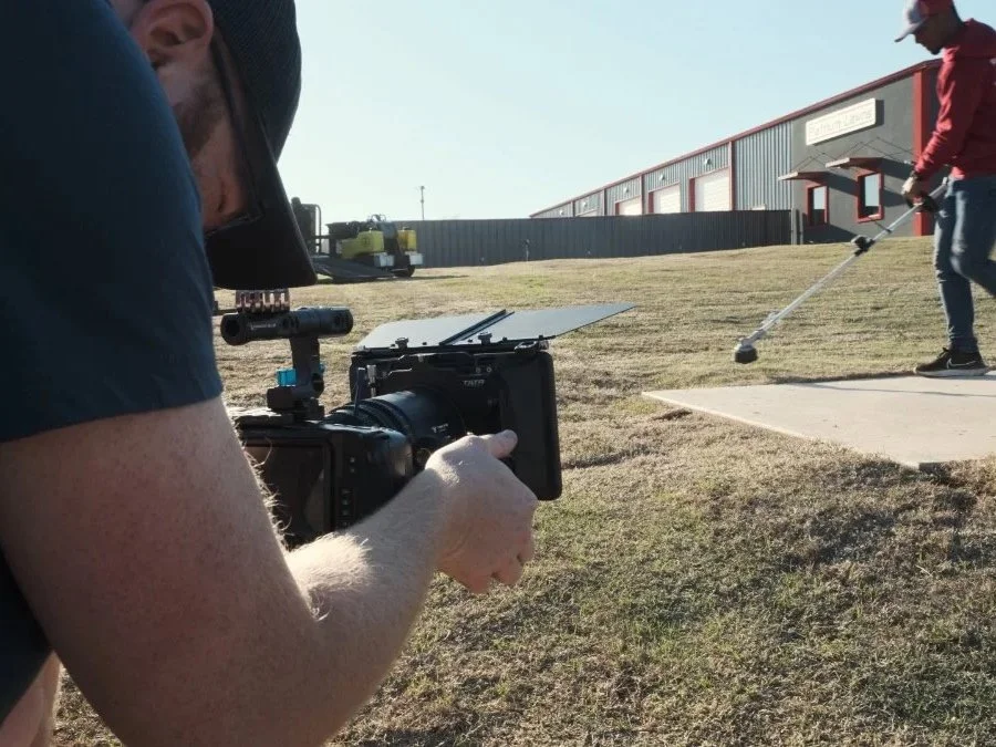 A person filming with a professional camera outdoors while another person is practicing golf with a golf club near a concrete mat.