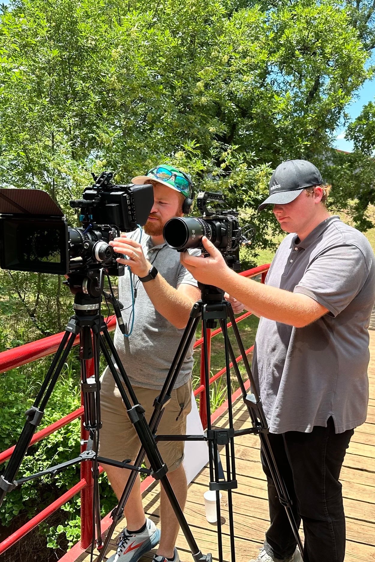 Two men filming outdoors on a wooden deck with trees in the background, using professional video cameras on tripods.