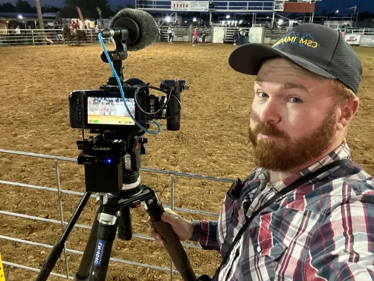 Man with a red beard and plaid shirt recording a rodeo event on a video camera at an outdoor arena during evening.