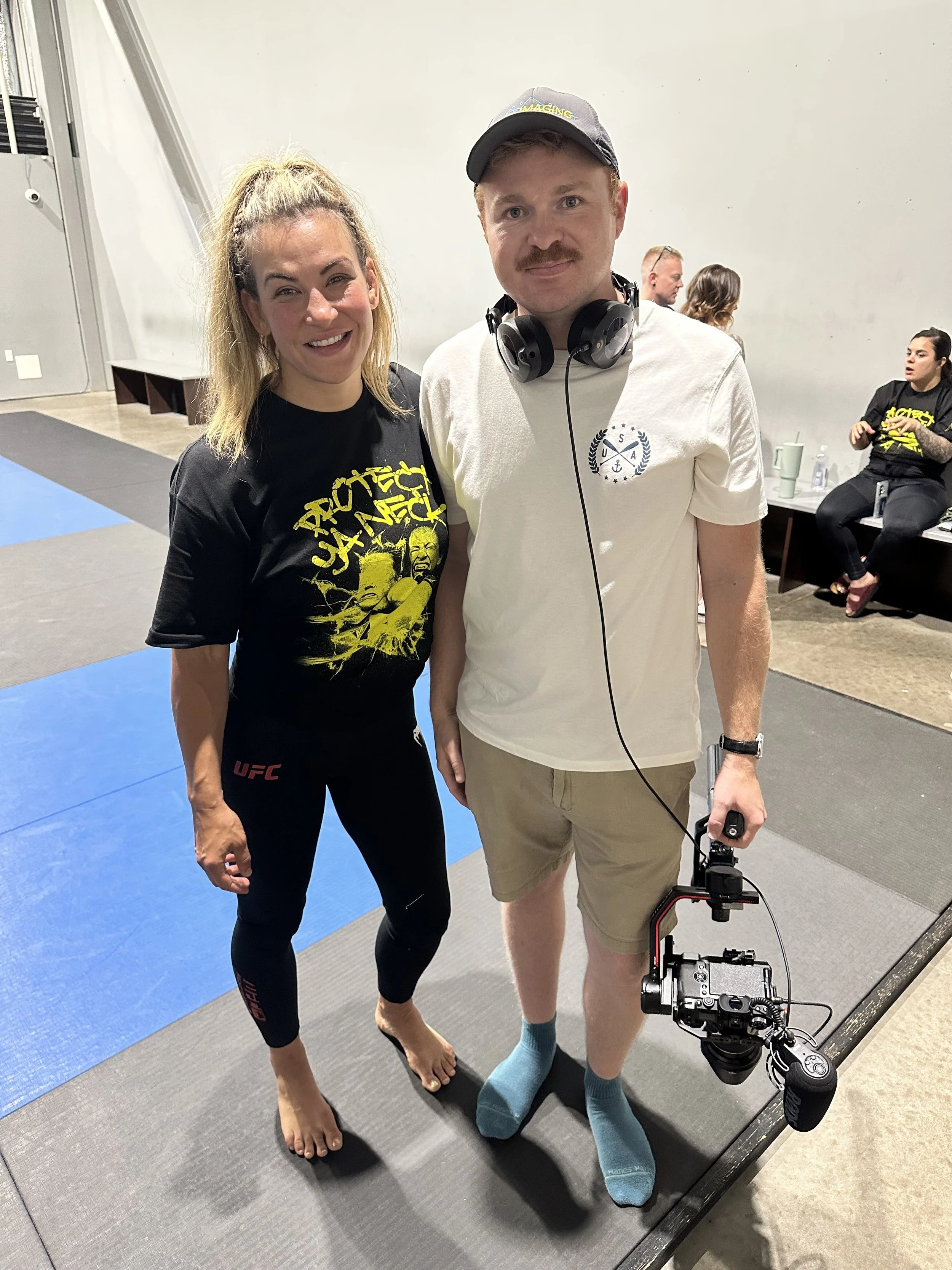 A woman in a black UFC T-shirt and black leggings standing barefoot on a mat, smiling next to a man in a white T-shirt, khaki shorts, and teal socks, holding a camera stabilized with a gimbal, in an indoor setting with a few people seated in the background.