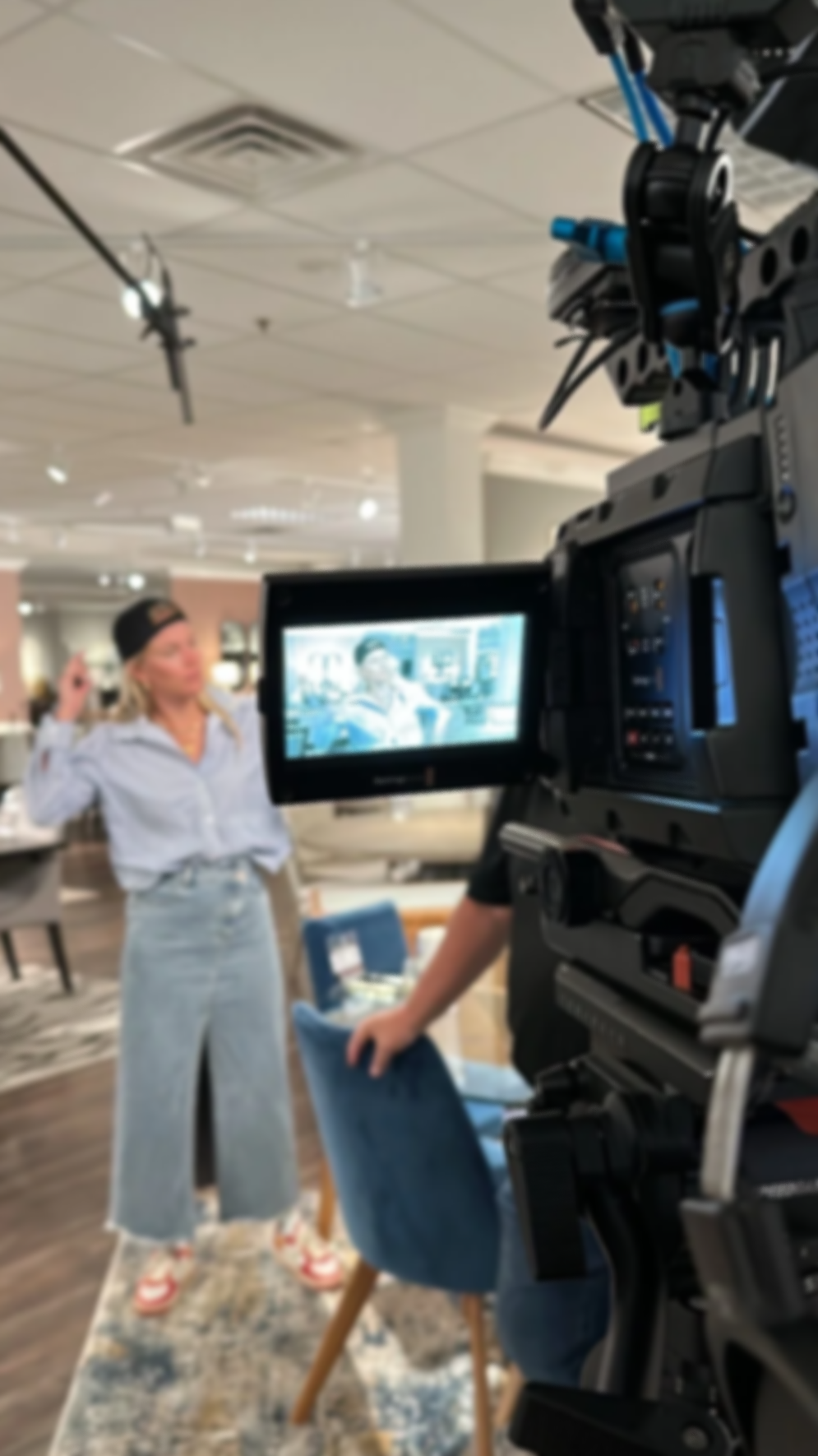 A woman on camera being interviewed or recorded in an indoor setting, with equipment and chairs visible around her.