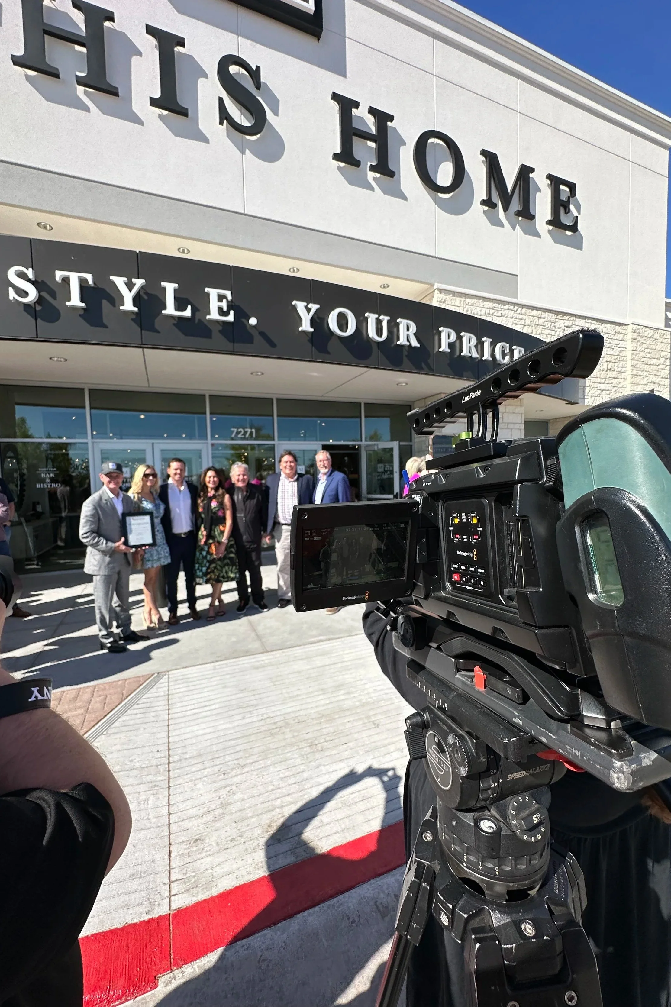 A group of people standing outside a retail store with a large sign reading 'Mathis Home' and a smaller sign displaying 'Style. Your Price.' A camera on a tripod records the scene.