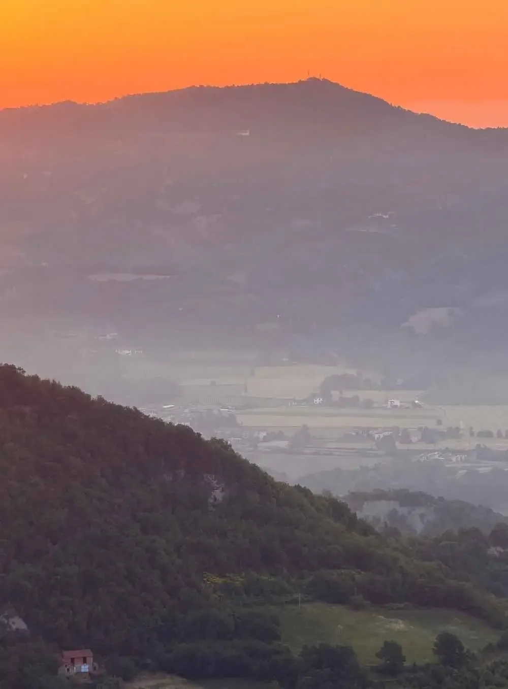 A distant mountain landscape during sunset with an orange sky, foggy valleys, and lush green hills in the foreground.