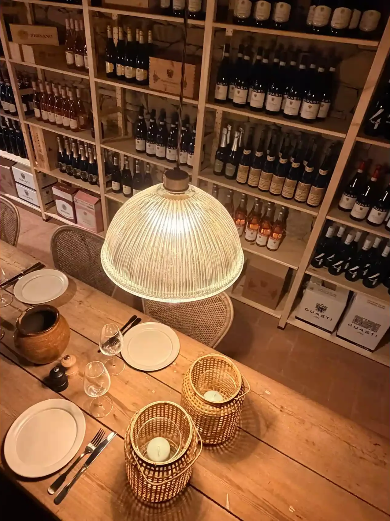 Wooden dining table set with white plates, silverware, glasses, and woven candles in baskets. A large pendant light hangs over the table. Behind, shelves hold wine bottles and boxes in a cozy, rustic restaurant setting.