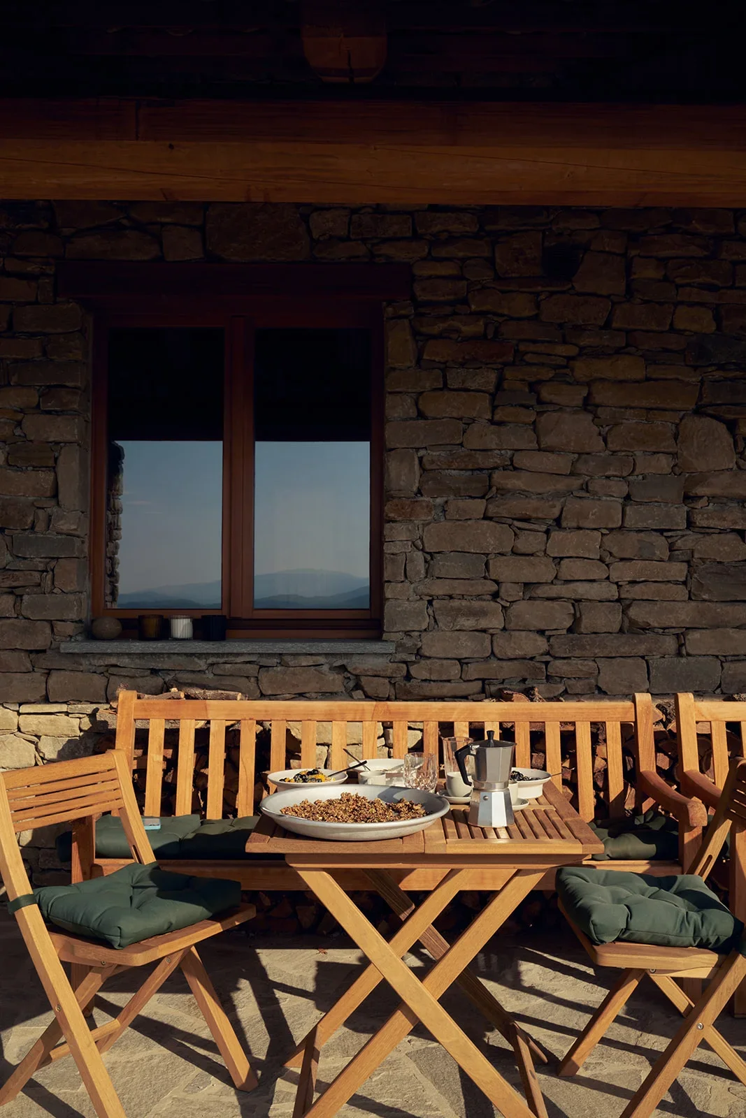 An outdoor wooden table set with food and drinks, surrounded by wooden chairs with cushions, against a stone wall with a window showing mountainous scenery.