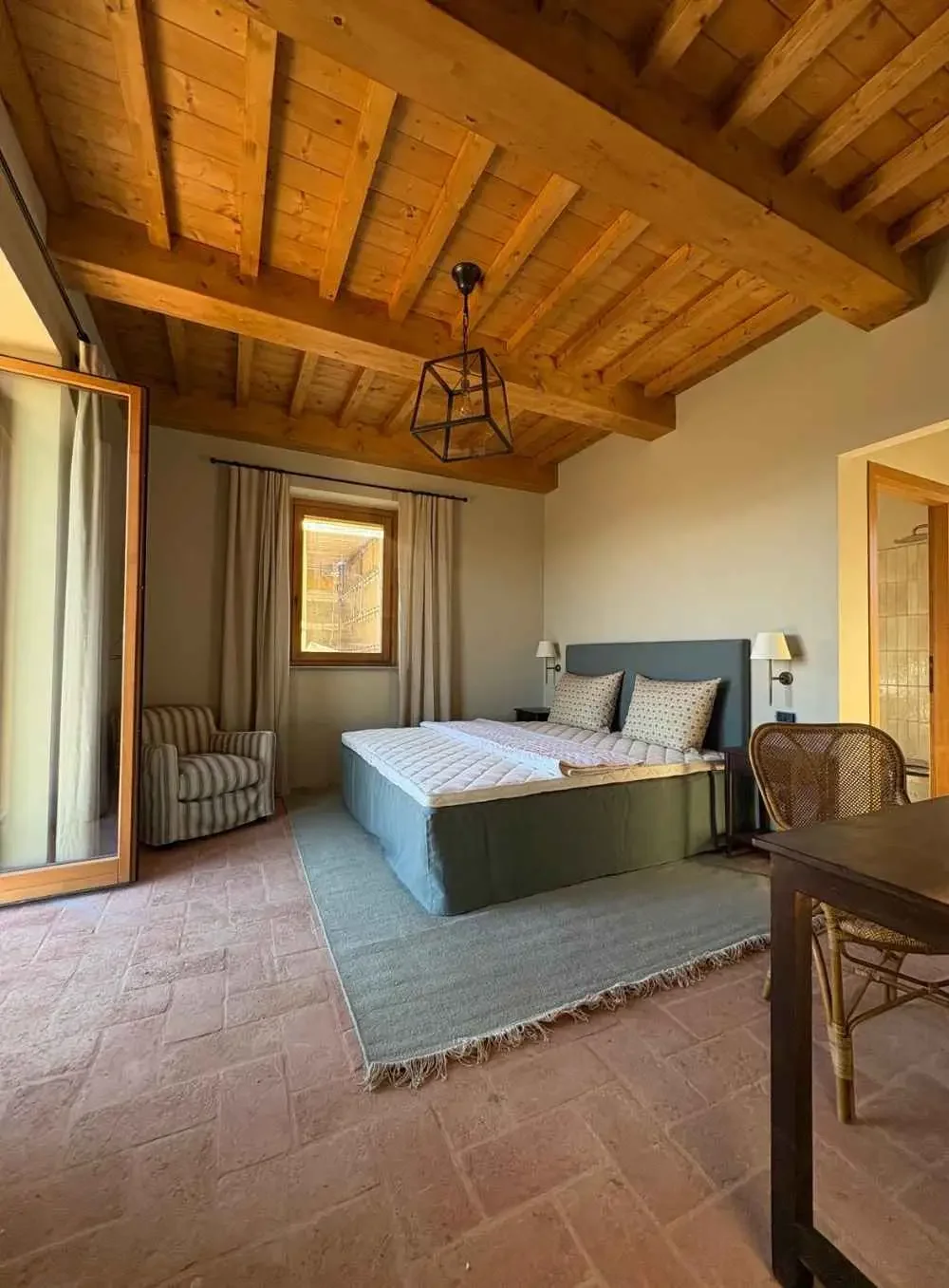 A cozy bedroom with a wooden ceiling, a bed with a blue headboard, a small armchair with beige and white stripes near a window, and a wicker chair at a table.