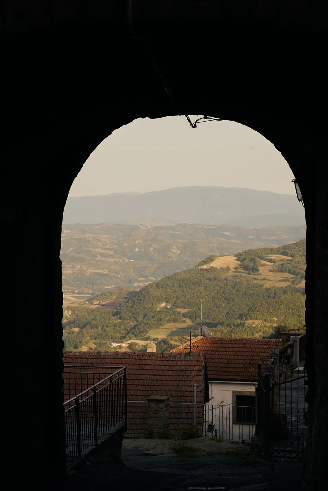 Mountain landscape viewed through a stone archway with rooftops in the foreground.