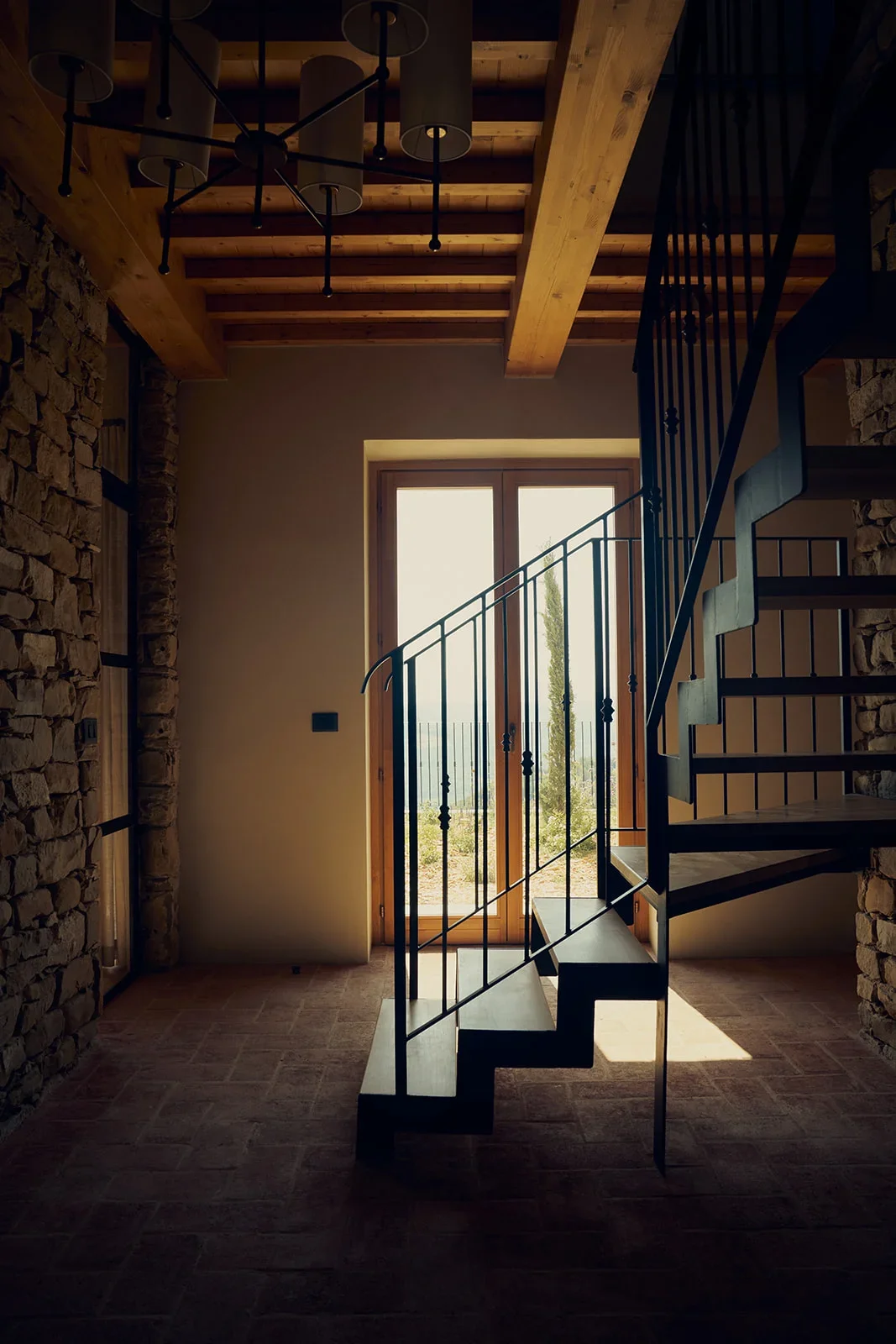 Staircase inside a rustic home with stone walls, wooden ceiling beams, large glass door letting in natural light, and a modern black metal spiral staircase.