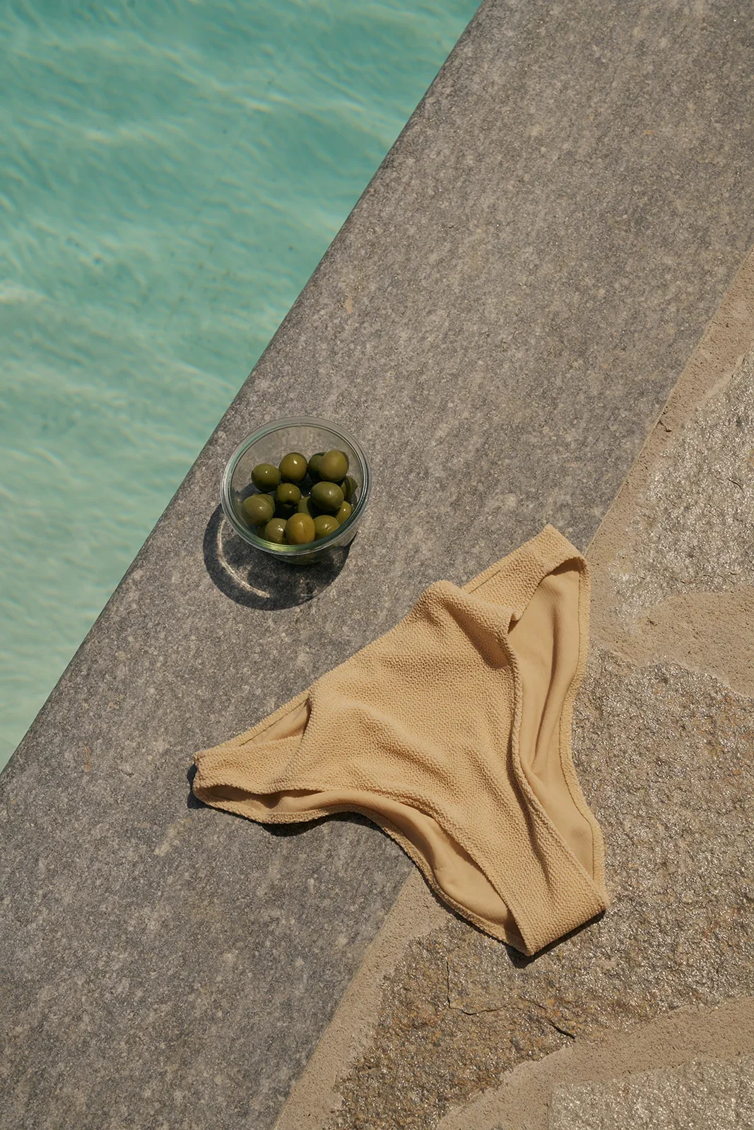 A clear glass bowl of green olives on a poolside ledge, next to a beige swim bottom and a towel, with a swimming pool in the background.