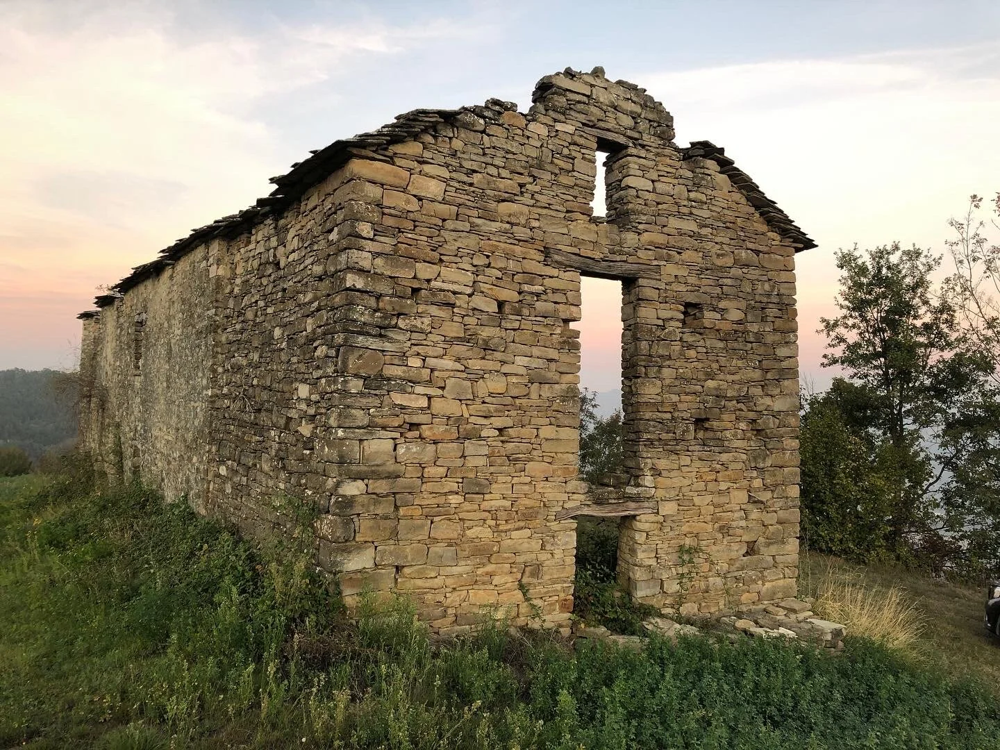 Old stone building ruins with missing roof, surrounded by grass and trees, during sunset or sunrise.