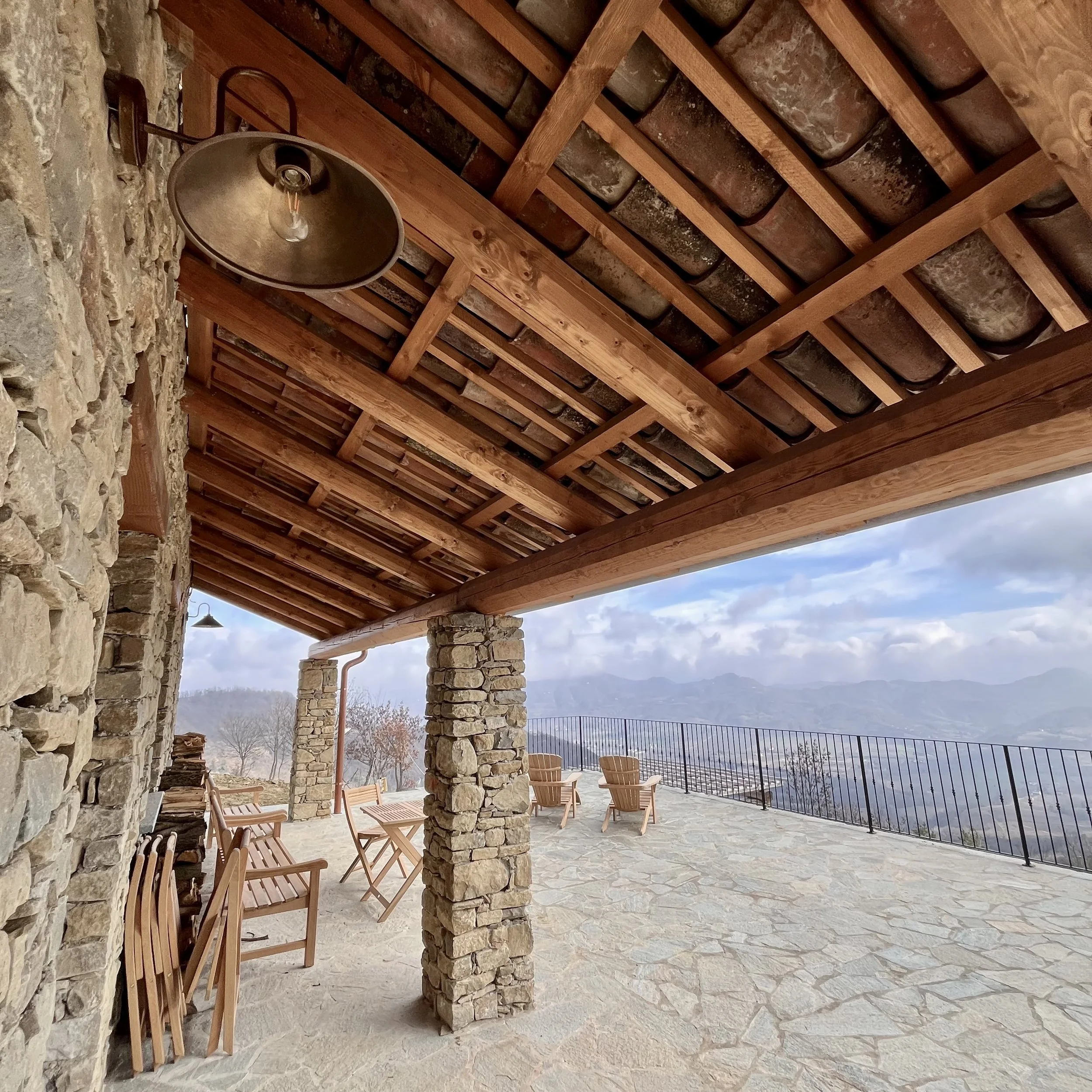 An outdoor stone patio with wooden chairs and tables, supported by stone columns and a wooden roof, overlooking a mountain landscape with cloudy skies.