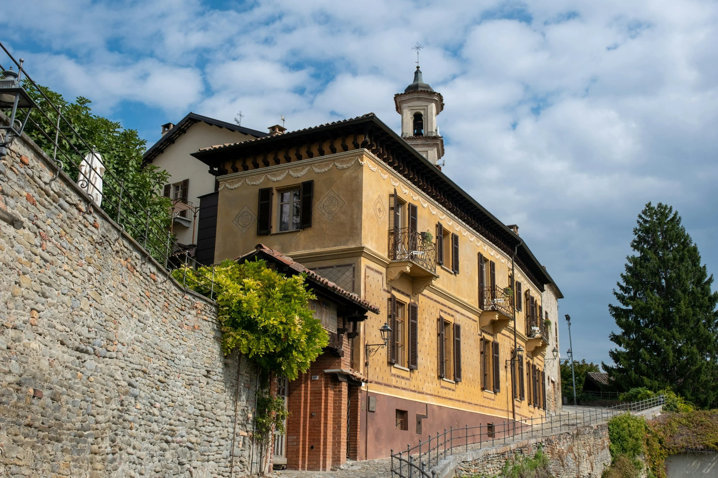 A historic yellow and beige building with dark brown shutters and small balconies, situated on a hillside with a stone retaining wall and a cobblestone path, under a partly cloudy sky with a tall green tree nearby.