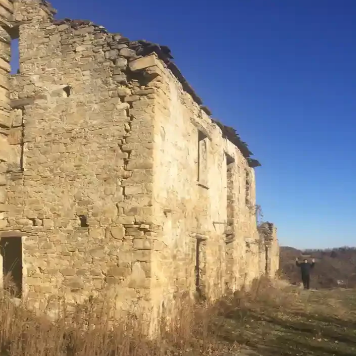 An old stone building with a damaged roof and missing windows, standing on a grassy landscape under a clear blue sky, with a person in the distance.