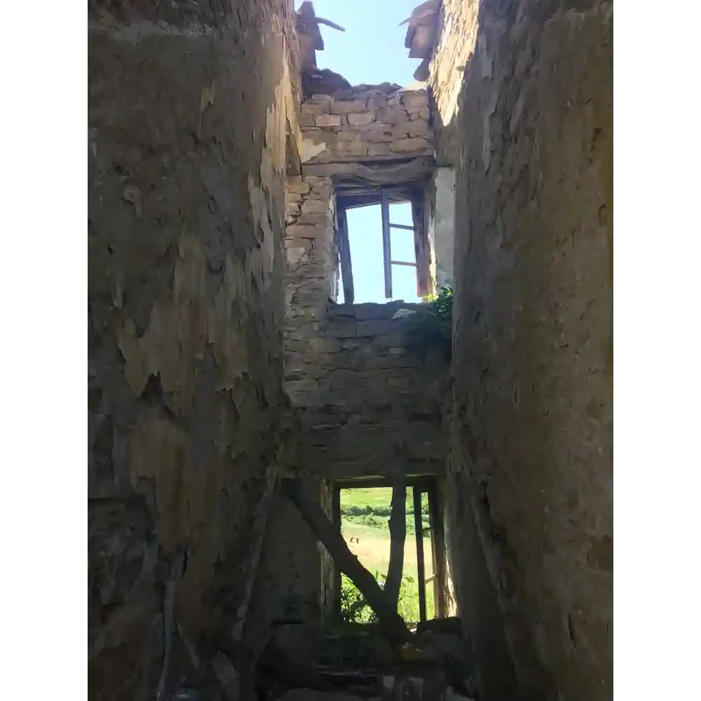 View of the interior of an abandoned stone building with two rectangular windows, showing a glimpse of the sky and greenery outside.