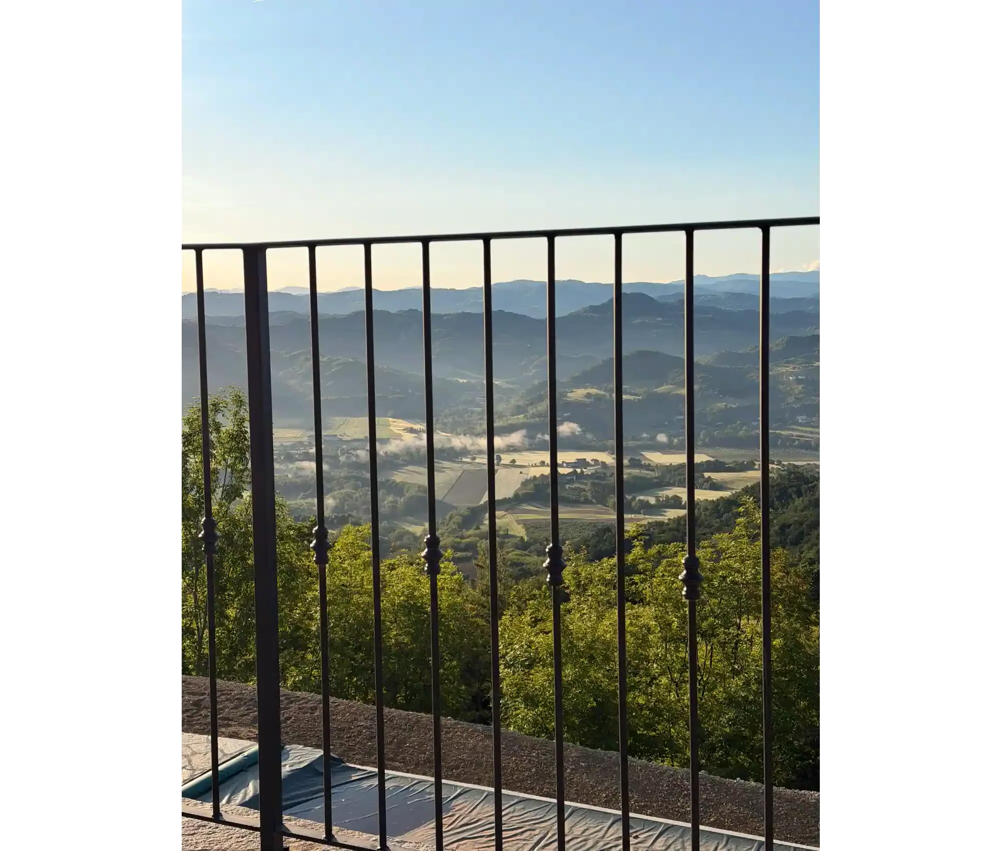 View of mountains and valley seen from a balcony with black railing, trees in the foreground, blue sky and some fog at the valley level.