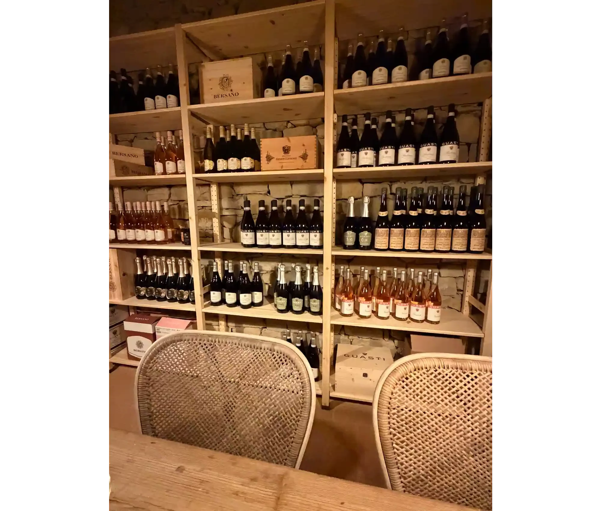 A wine cellar with wooden shelves holding multiple bottles of wine and crates. There are two wicker chairs in the foreground.
