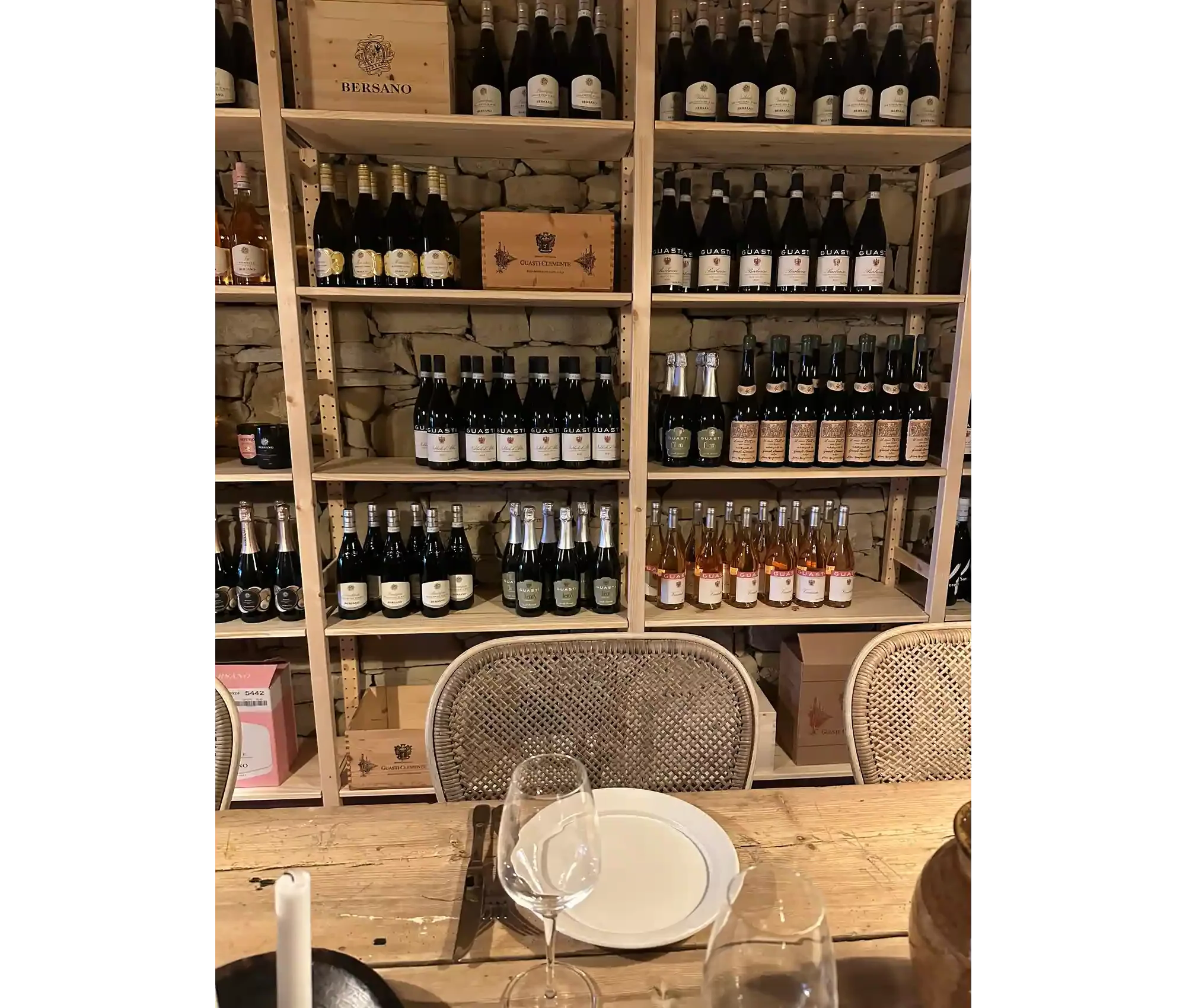 A wooden shelving unit filled with various bottles of wine or sparkling wine, with some label details visible. The shelves are against a stone wall, and there are chairs and a table set with a wine glass and a plate in the foreground.