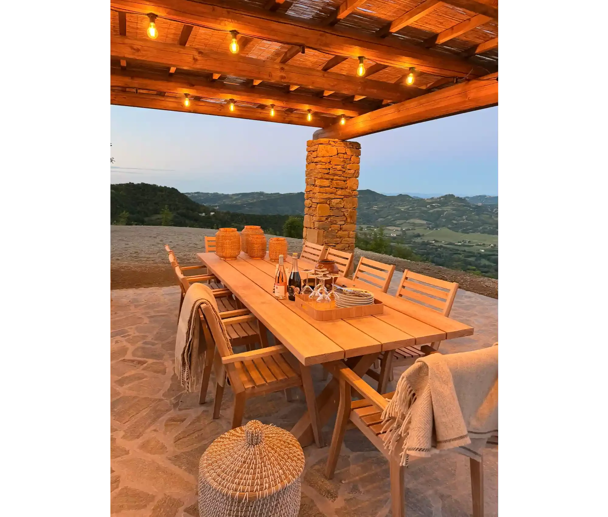 Outdoor dining area with a long wooden table set for a meal, surrounded by wooden chairs, and decorated with woven baskets and bottles. The area is covered by a wooden pergola with string lights, and overlooking a landscape of rolling hills and blue 