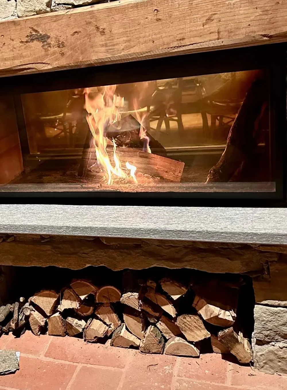 A close-up view of a modern fireplace with a glass front, showing burning logs inside, with a neatly stacked pile of firewood underneath and a stone and wood mantle above.