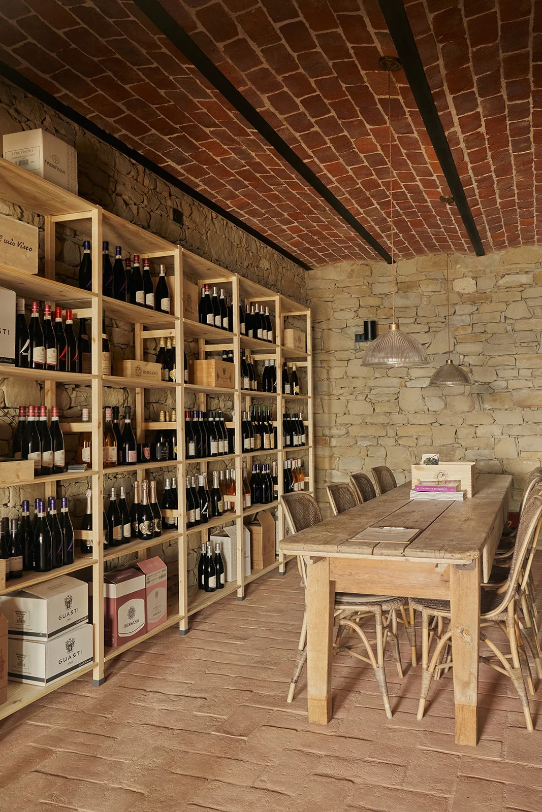 A cozy wine tasting room with brick ceiling, stone walls, wooden shelving filled with wine bottles, a wooden table with chairs, and hanging pendant lights.