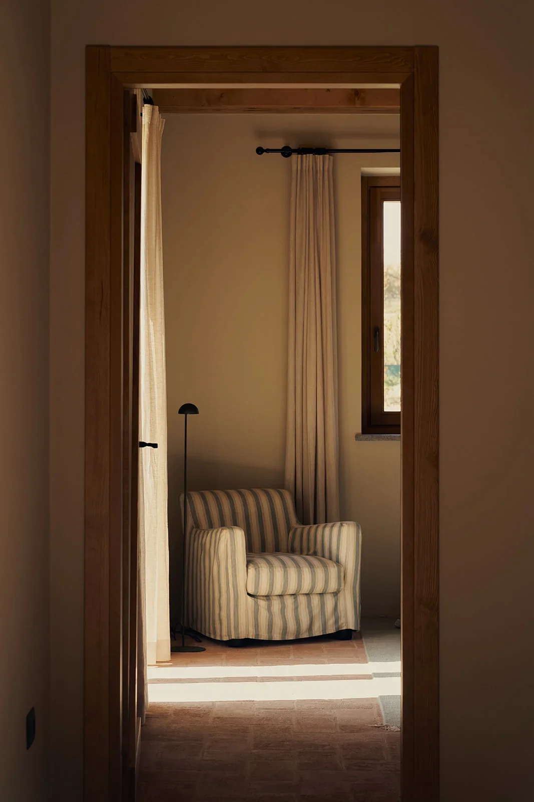 A cozy corner of a room with a striped armchair, a black floor lamp, light-colored curtains, and a window with natural light coming through.