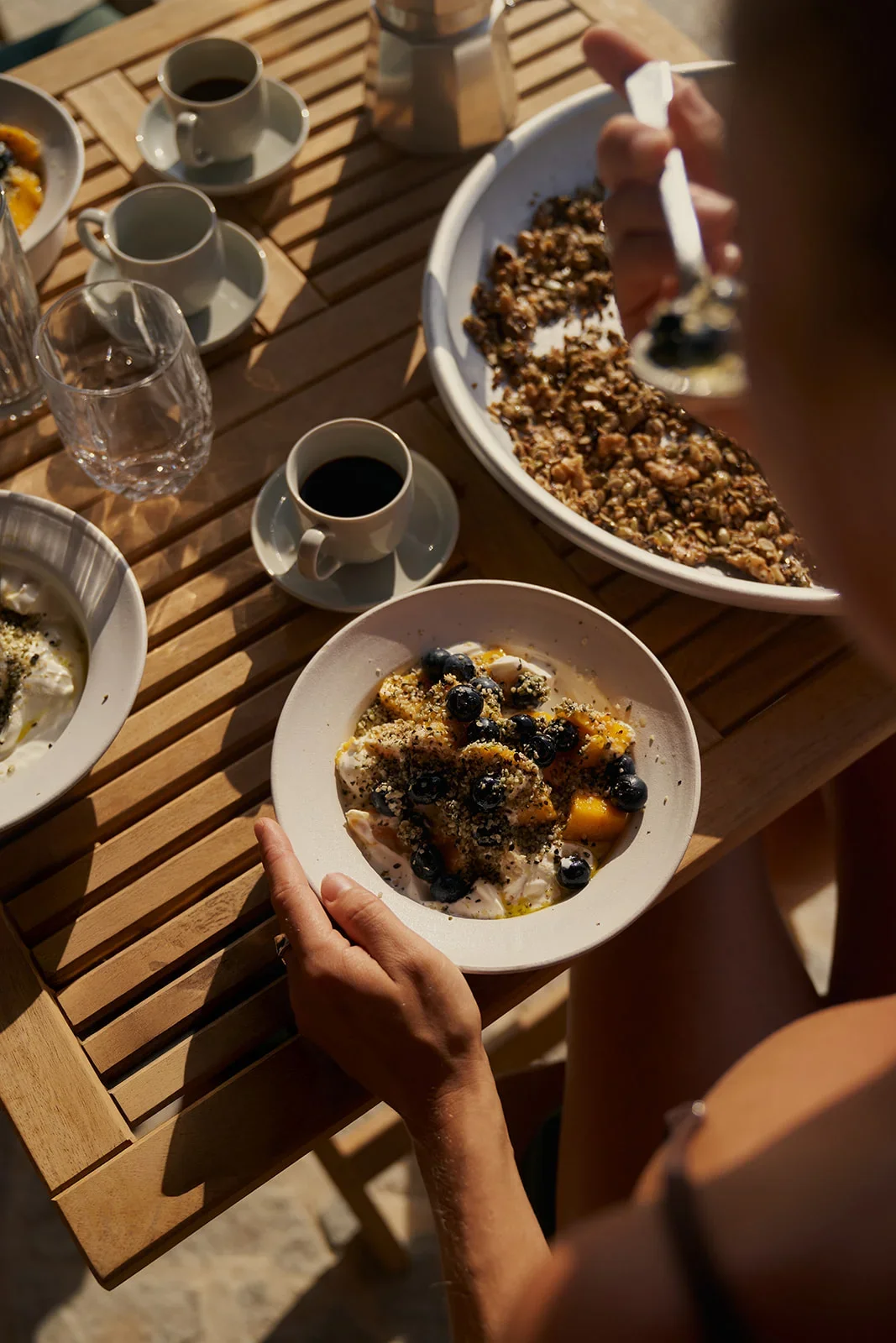 Person holding a bowl of yogurt topped with blueberries and fruit, with a table of coffee cups, plates, and desserts in the background.