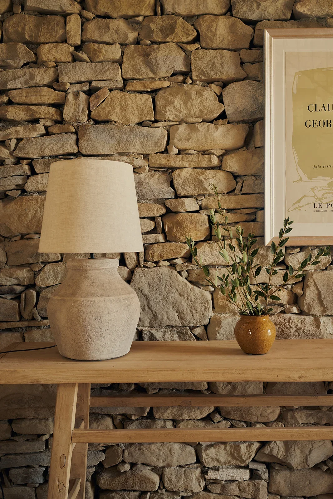 A rustic interior scene featuring a stone wall, a wooden shelf, a beige table lamp, and a small ceramic vase with green leafy branches, alongside a framed piece of art.