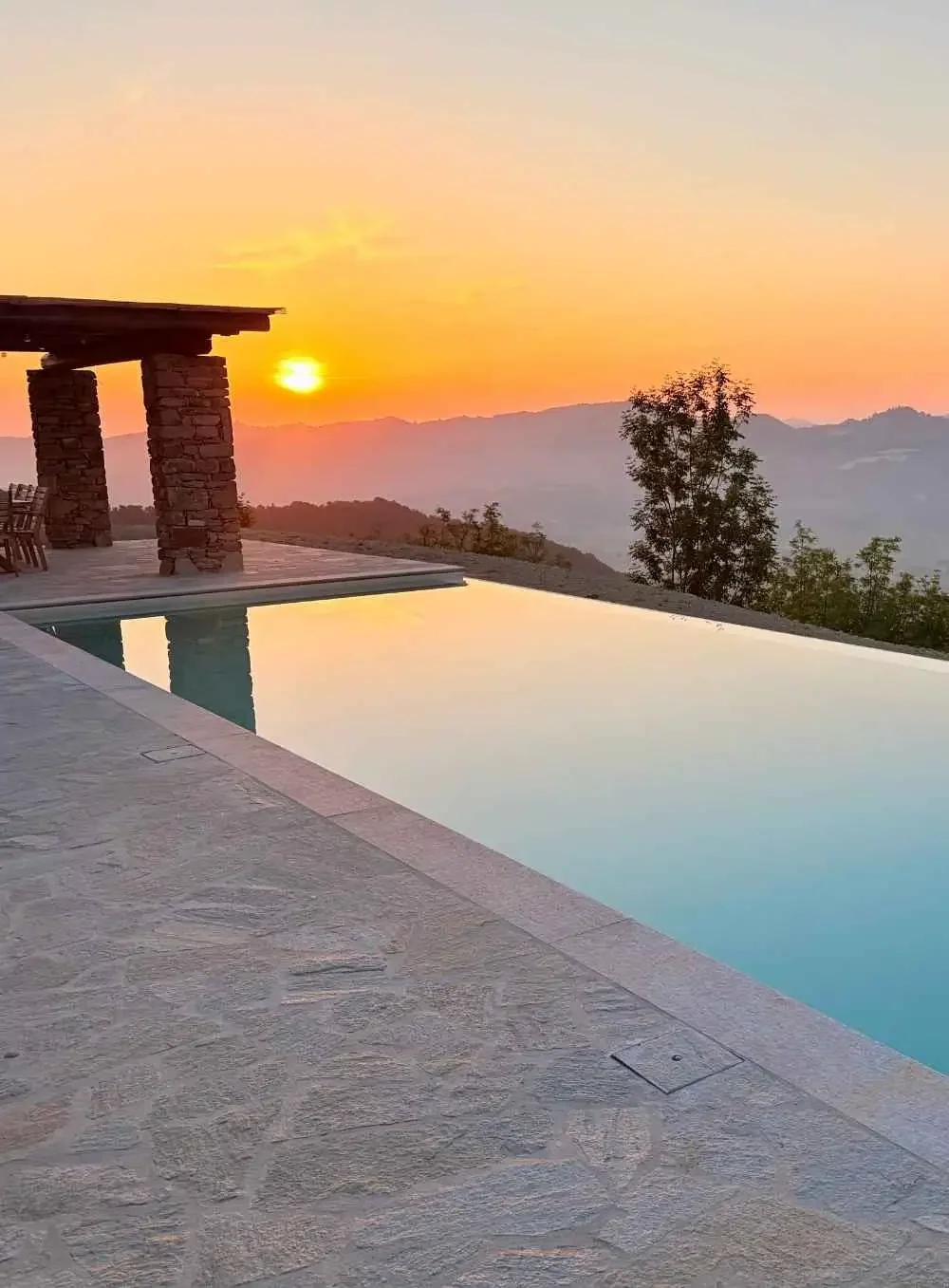 A picturesque sunset view over a mountain range, with an infinity pool reflecting the colorful sky in the foreground. There is a stone patio with outdoor furniture and a stone and wood pergola on the left side.