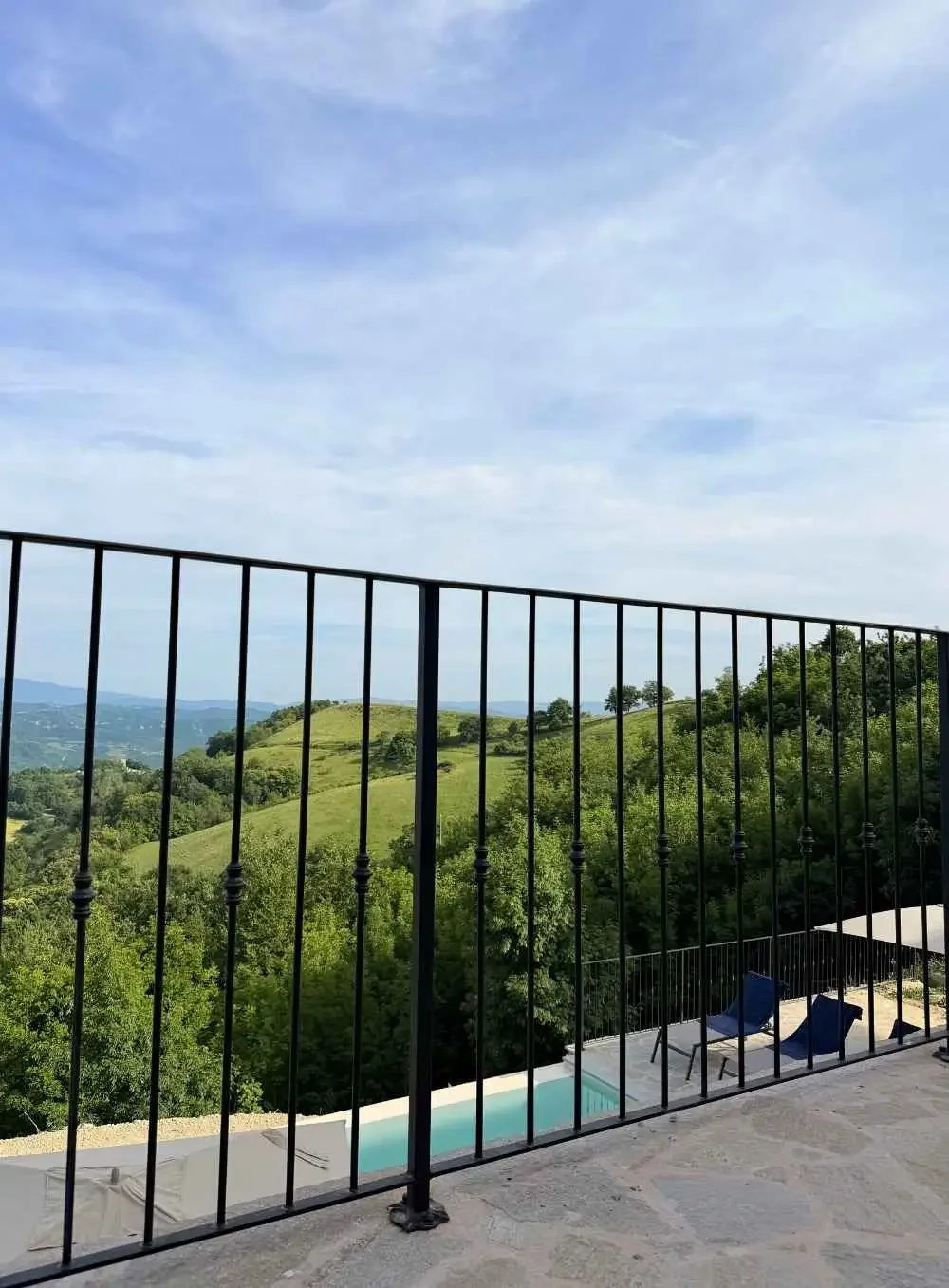 View of green rolling hills with trees, seen from a balcony with black metal railing; a small pool and two blue chairs are on the balcony.
