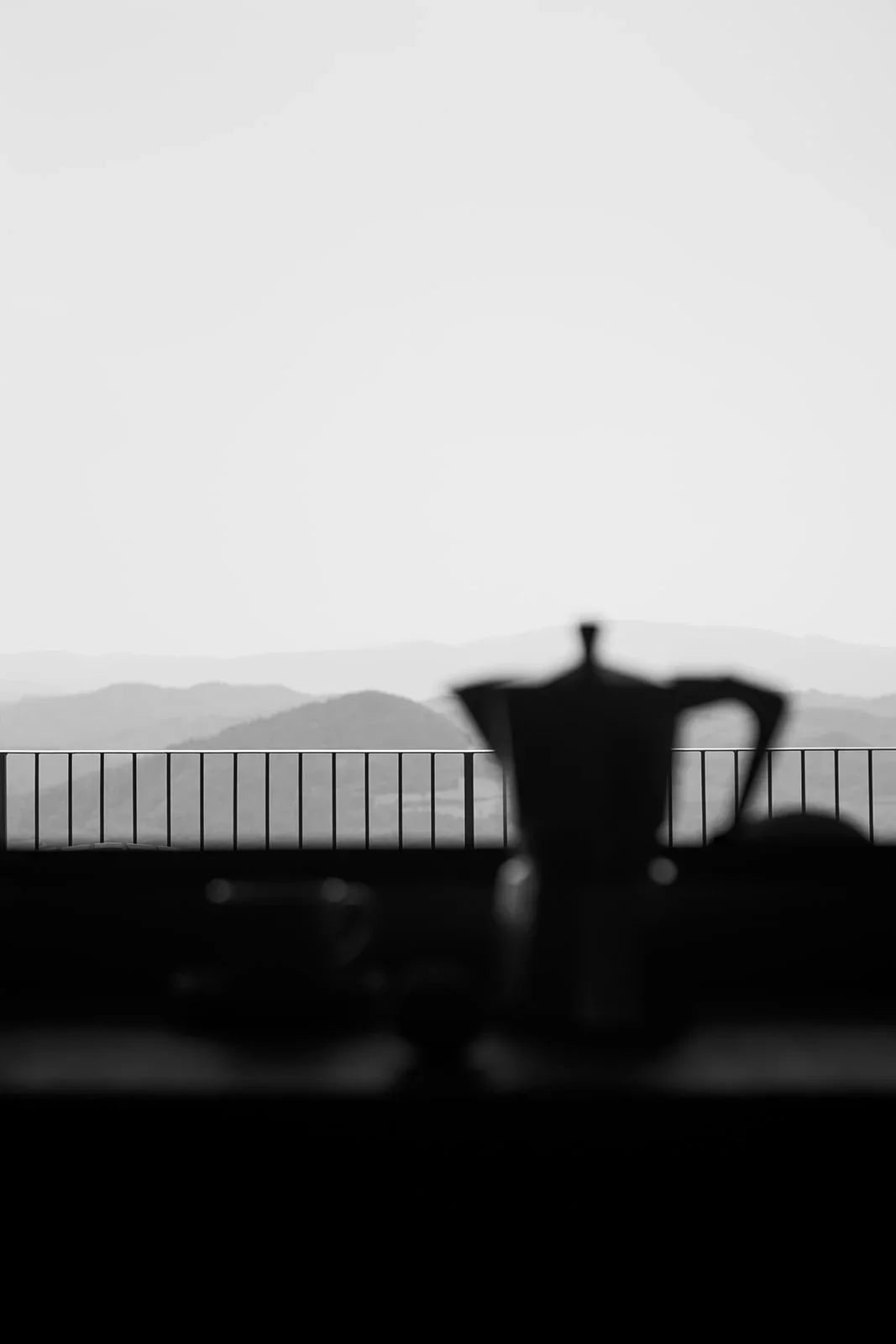 Silhouette of a coffee pot on a table with mountains in the background, seen on a balcony in black and white.